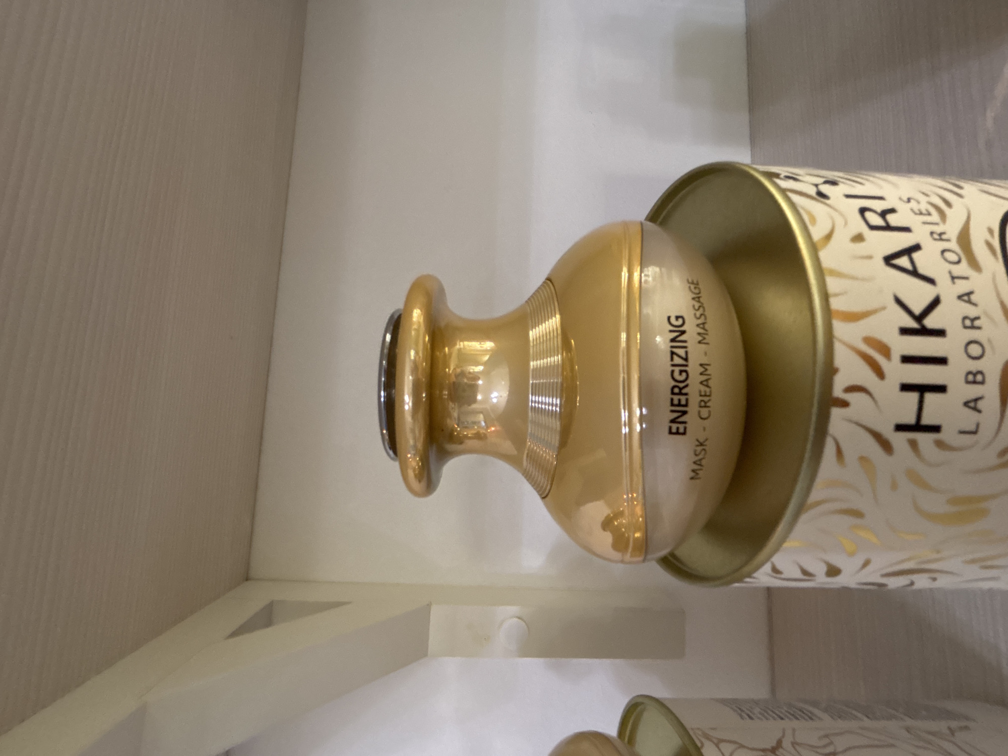 The image shows two products of Hikari Laboratories on a white shelf. The products appear to be face masks or creams, and are contained in golden cylindrical packaging with the "HIKARI LABORATORIES" logo printed on it. The word "ENERGIZING" is also visible on the packaging, along with "MASK - CREAM - MASSAGE". The products are oriented horizontally on the shelf, with one partially obscured by the other. The environment consists of a white shelf and wall. No specific location or city is identifiable.