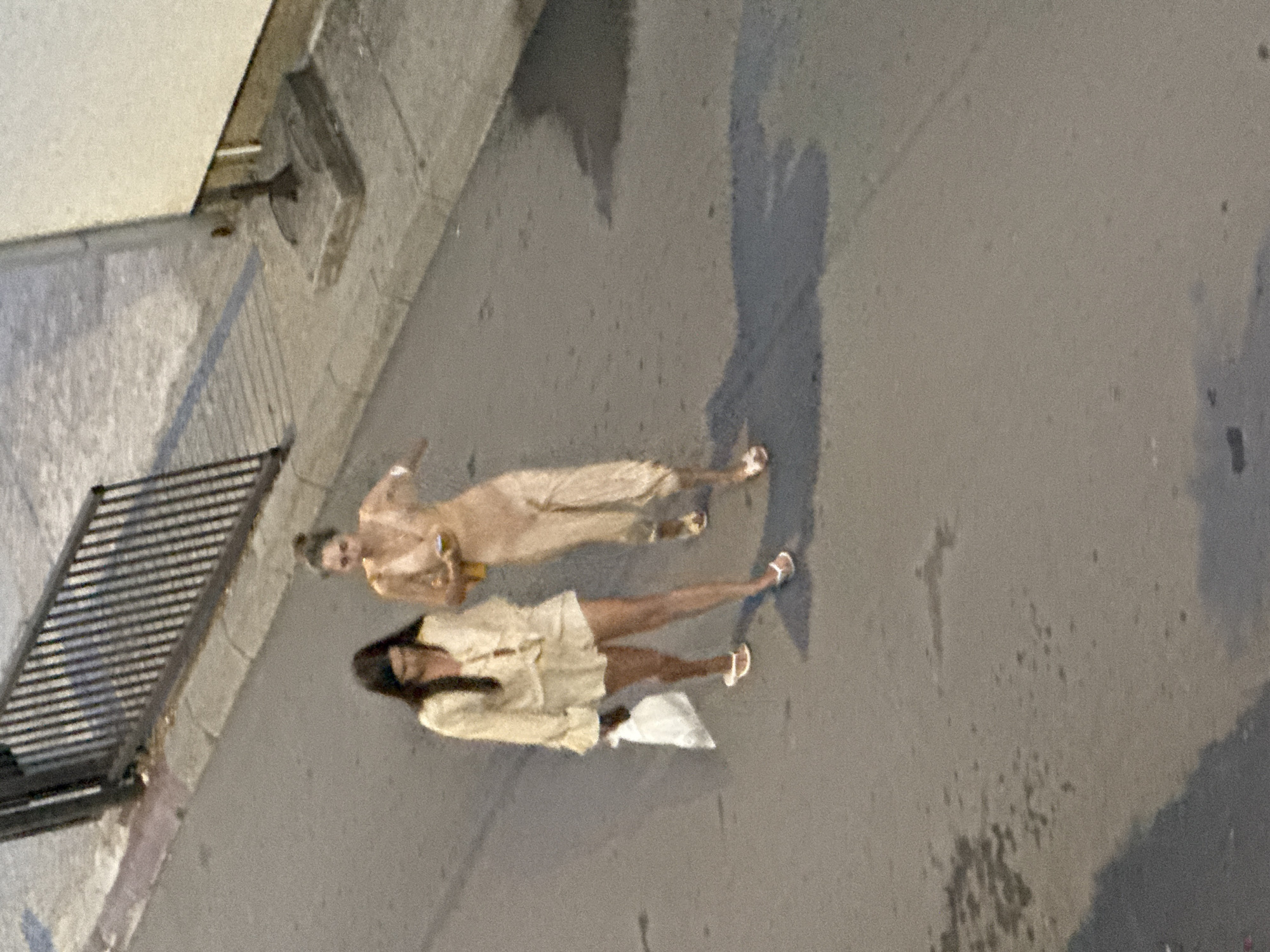 The image shows two women standing on a paved surface, likely a street or sidewalk, in Budva, Montenegro. The scene appears to be outdoors, and the lighting suggests it is either late afternoon or early evening, with long shadows cast on the ground. One woman, in the foreground, is wearing a light-colored, short-sleeved top and a skirt, with white sandals. She is carrying a white bag. Her hair is dark and appears to be pulled back. The second woman, standing behind and to the left of the first, is wearing a long, light-colored, possibly satin dress and matching sandals. She is holding a drink in her right hand. Both women are viewed from above, indicating the photo was taken from an elevated position. A dark metal fence is visible on the left side of the image, and a textured wall or building is present behind it. The ground is a speckled gray pavement. No text is visible in the image.