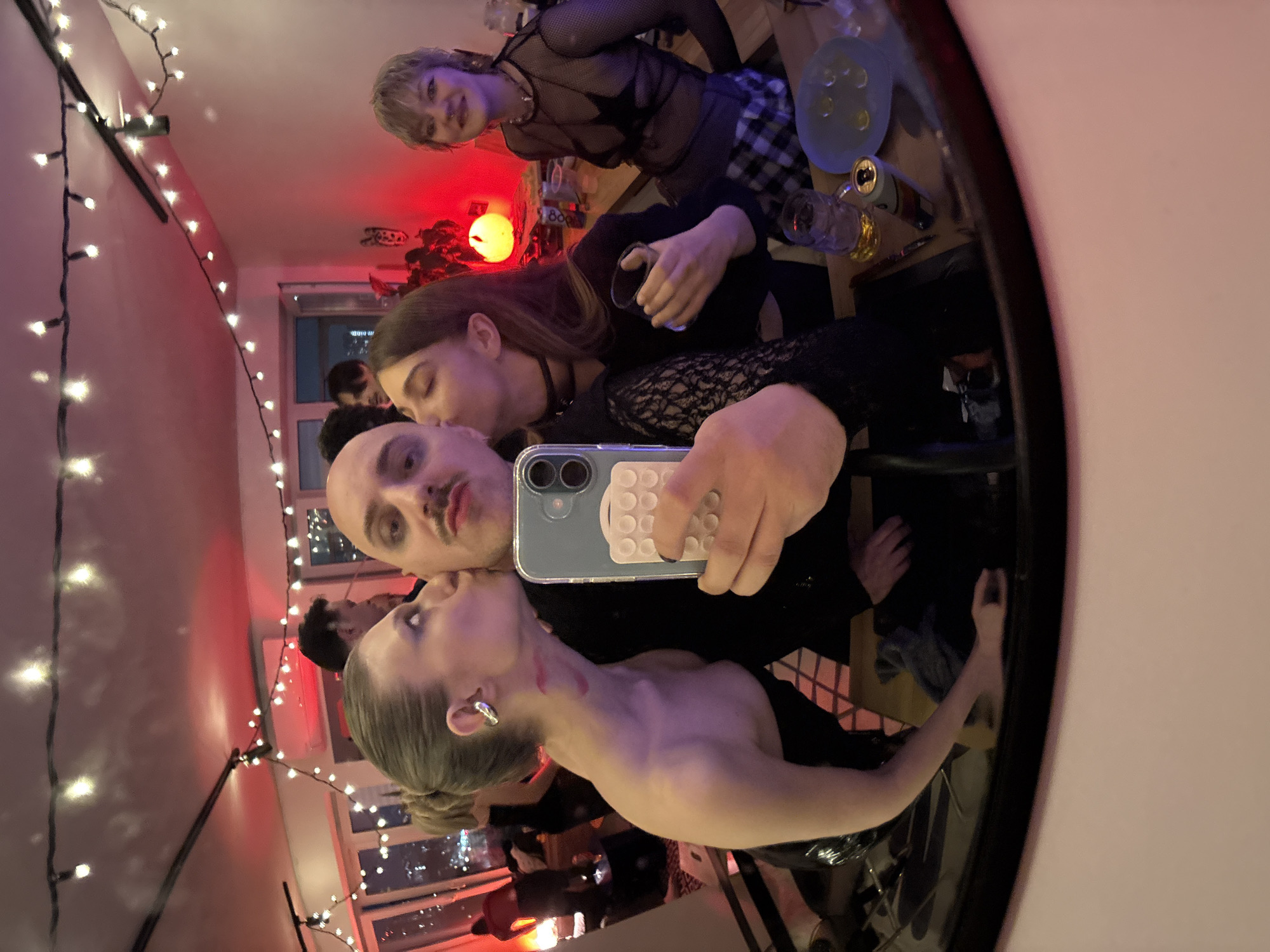 A reflective view captures a group of individuals in an indoor setting. Three people appear prominently: a woman with long brown hair kisses the cheek of a bald man who is holding up a phone. A second woman with gray-blond hair is nuzzled close to the bald man. These three people are the apparent subjects of the photo, and the bald man is holding the smartphone up to take the picture. To the upper right, in the reflection is seen a woman wearing a black mesh top and a plaid skirt. Behind the three, more figures stand with a view of the city buildings outside a window. String lights hang from the ceiling, and some additional lighting features are visible in the room's interior, as well as food and drink items. A mirror frame encompasses the view. The setting is identified as Moscow, Russia, based on the urban skyline view.