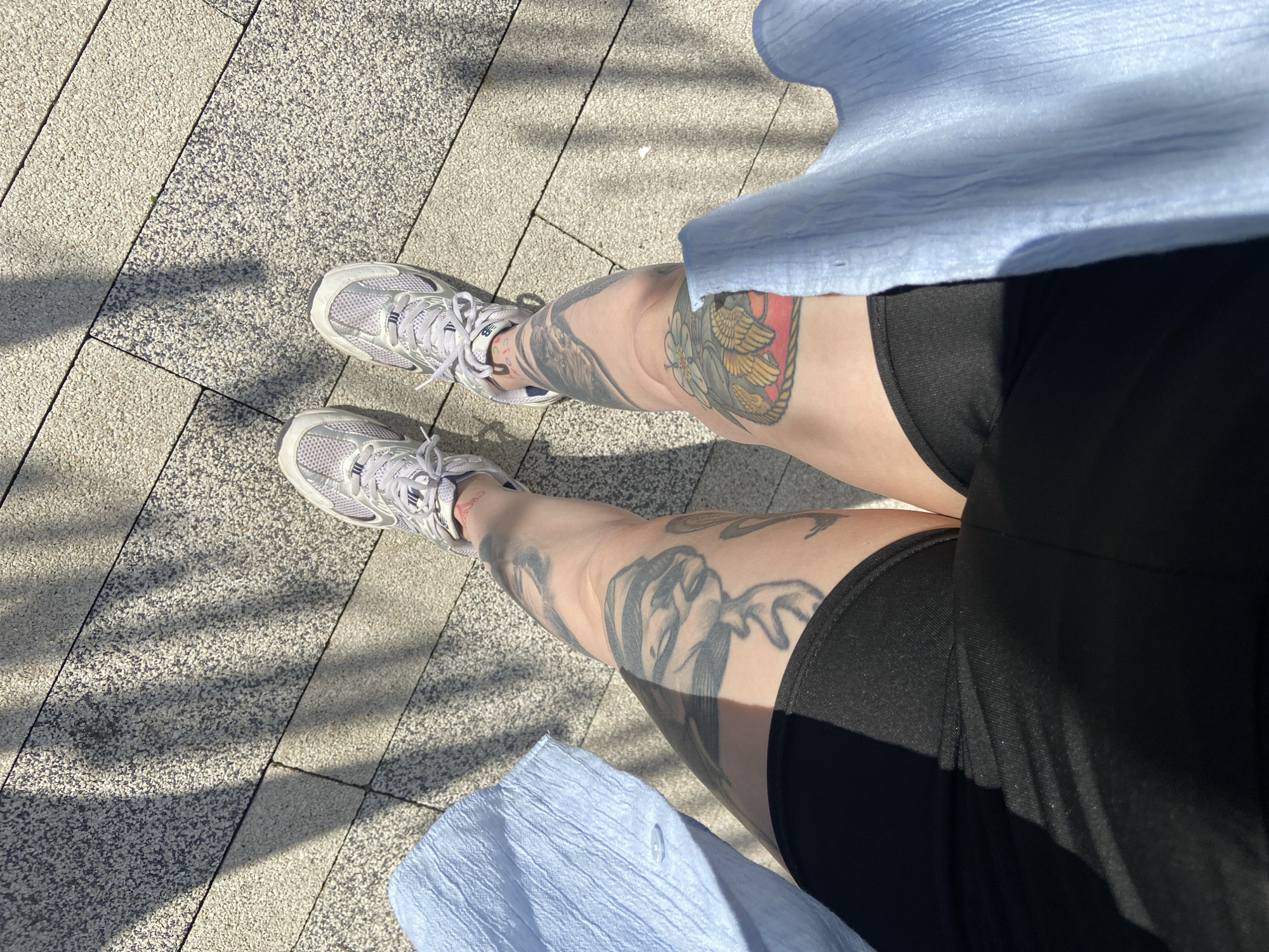 The image shows a person sitting outdoors on a paved surface of grey and white bricks with dark lines. The person is wearing black shorts, a light blue shirt, and grey athletic shoes. Both legs are visible and covered in tattoos. Shadows from trees or foliage are visible across the pavement. The location in the city of Saratov, Russia, is not discernible from this image.