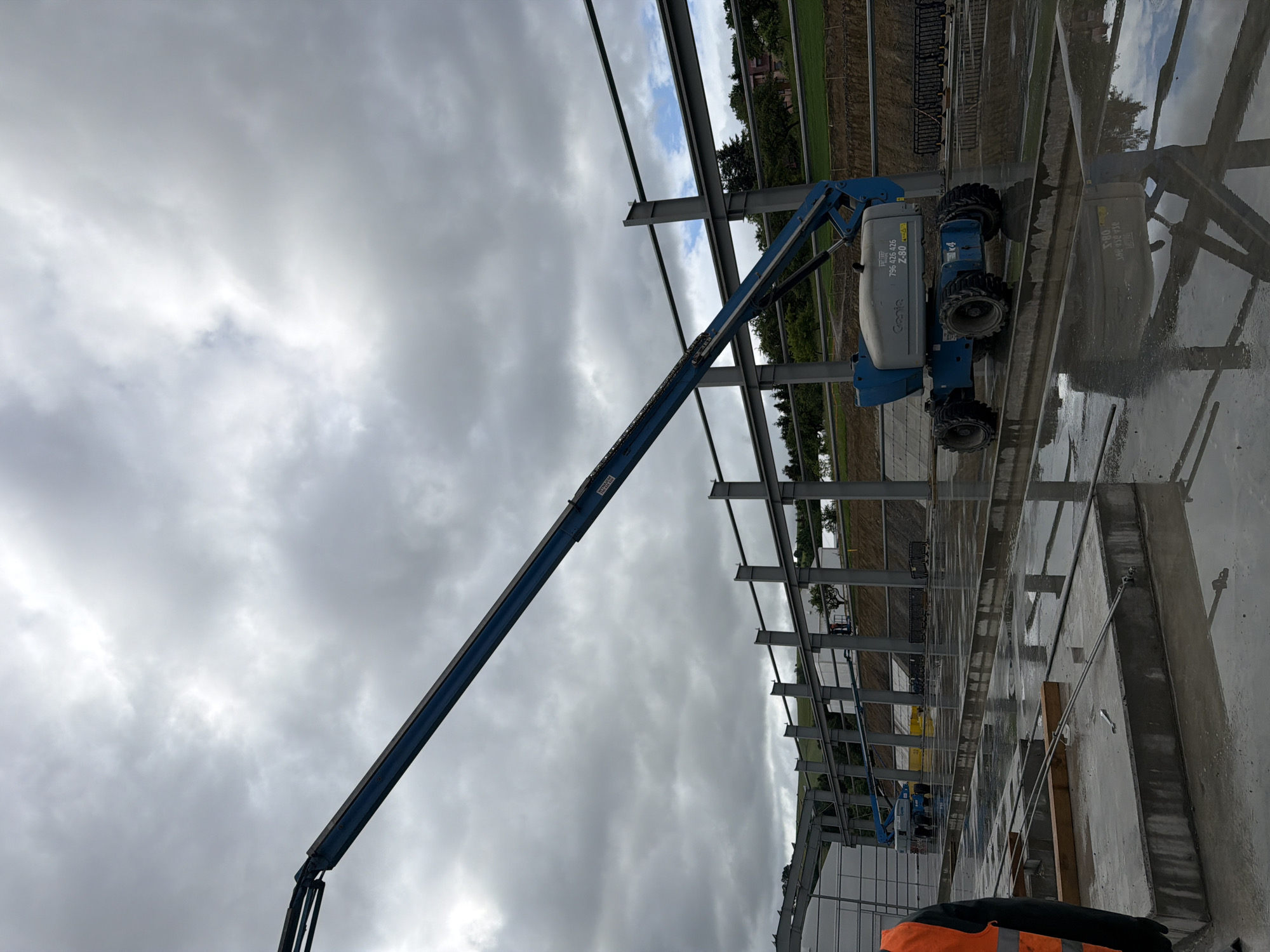 This is an outdoor, slightly elevated, full shot of a construction site with a metallic building frame under construction. The sky is heavily overcast with dramatic grey clouds. In the foreground, a person wearing an orange safety vest and a dark shirt is visible from the chest down on the left side of the frame. In the mid-ground, a blue Genie Z-80 articulated boom lift is positioned on a wet, reflective concrete surface. The boom lift has four large, off-road tires and its arm is extended upwards and to the left, over the construction site. The skeletal structure of a building is visible, consisting of numerous vertical steel posts and horizontal beams. The ground is wet and shows reflections of the construction elements. The background features a grassy hillside with scattered trees and some buildings. The image appears to be taken during the daytime.