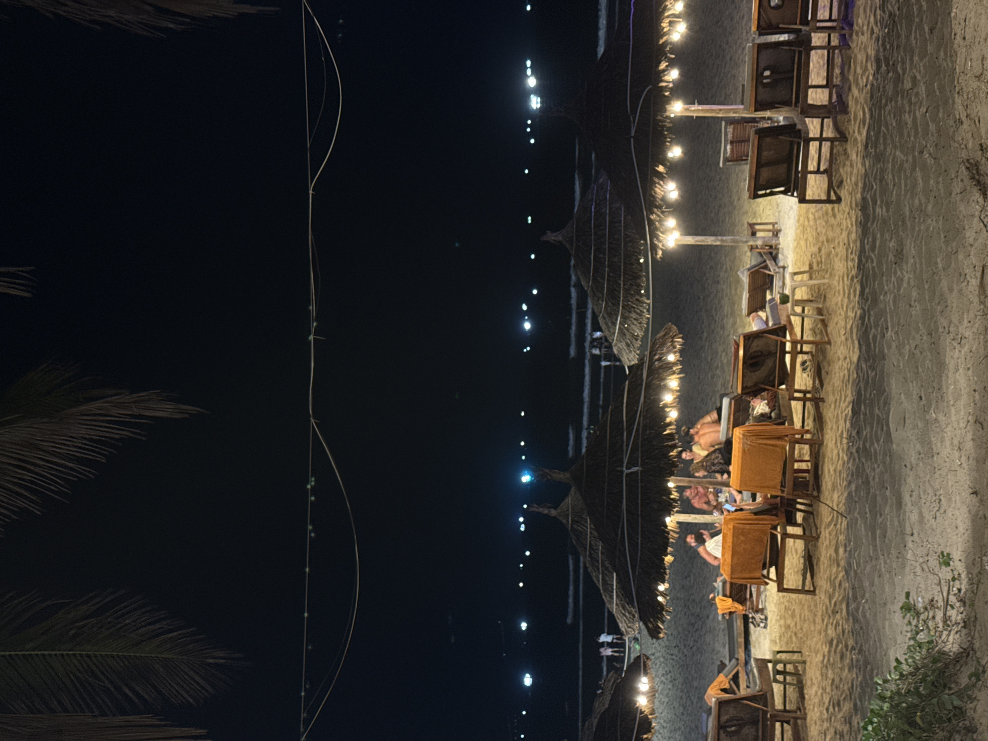 This media file captures a nighttime scene at a beachside establishment, likely a restaurant or bar, in Đà Nẵng, Vietnam. The image is oriented vertically, but when viewed horizontally, it shows a sandy beach on the right with the ocean and dark sky on the left.
The setting features rows of wooden tables and chairs placed directly on the sand. Many of the tables are covered with orange or tan tablecloths. Overhead, large, thatched umbrellas provide cover. The area is illuminated by numerous string lights strung between poles and umbrellas, casting a warm, inviting glow on the dining space.
Several people are visible, seated at the tables, appearing to be dining or socializing. Their faces are not clearly discernible due to the low light and distance, but their presence indicates an active, lively atmosphere.
On the far left (when viewed horizontally), the dark expanse of the ocean is visible, punctuated by a series of small, bright lights on the distant horizon, possibly from boats or a far-off coastline. Silhouetted against the dark sky on the bottom left are the fronds of a palm tree, adding to the tropical ambiance. Thin wires or ropes are also faintly visible stretching across the dark sky. The overall impression is one of a relaxed and pleasant evening spent by the sea. No text is visible in the image.