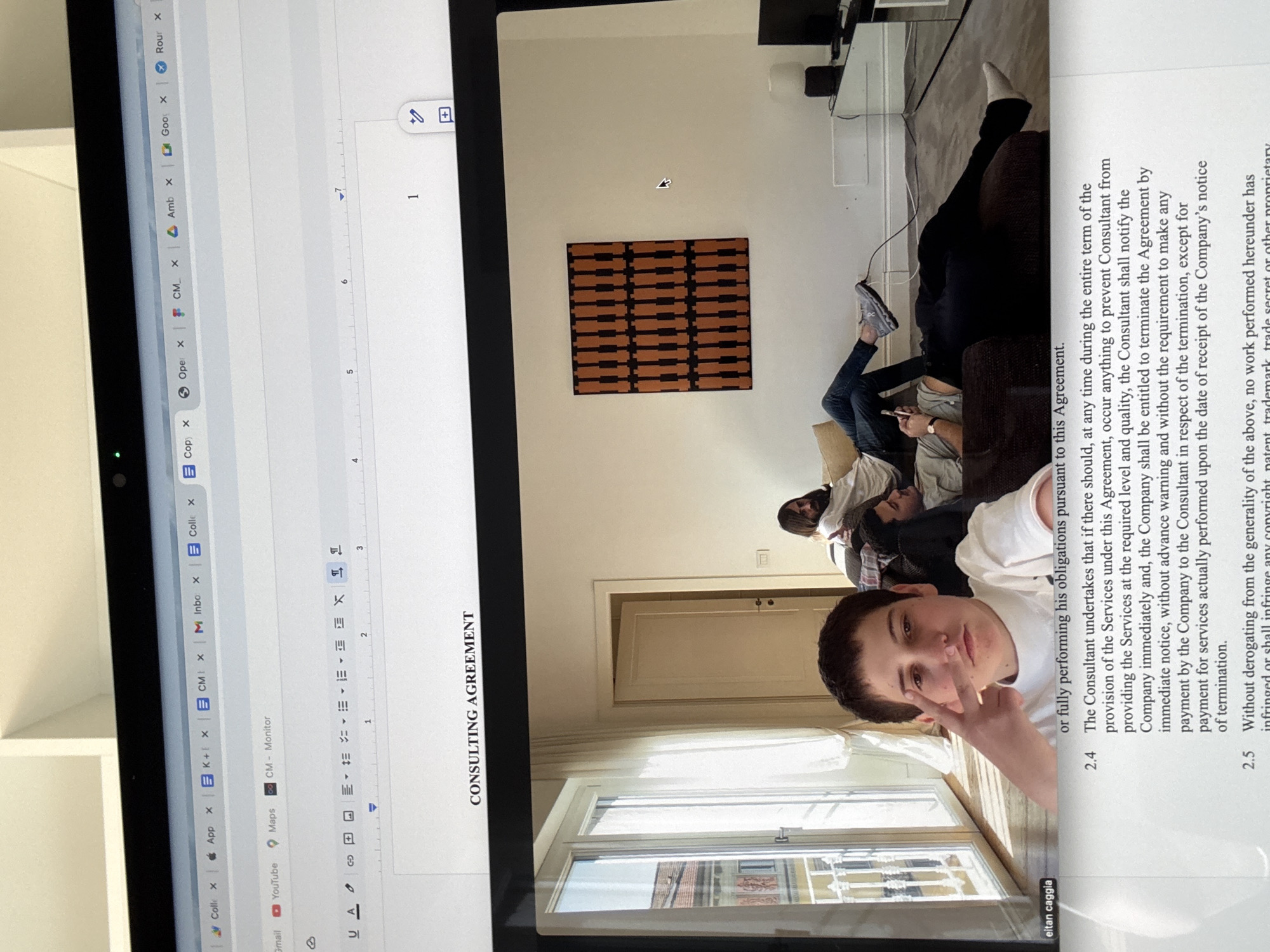 The image depicts a laptop screen showing two separate windows side-by-side. The left window displays multiple browser tabs. The right window displays a partially visible consulting agreement in a word processor, with the title "CONSULTING AGREEMENT" visible. A selfie of a young male partly obscures the text of the document.
The background visible through the window in the photograph seems to be the city of Milan, Italy. In the background of the image are three people reclining on a couch and a piece of artwork.