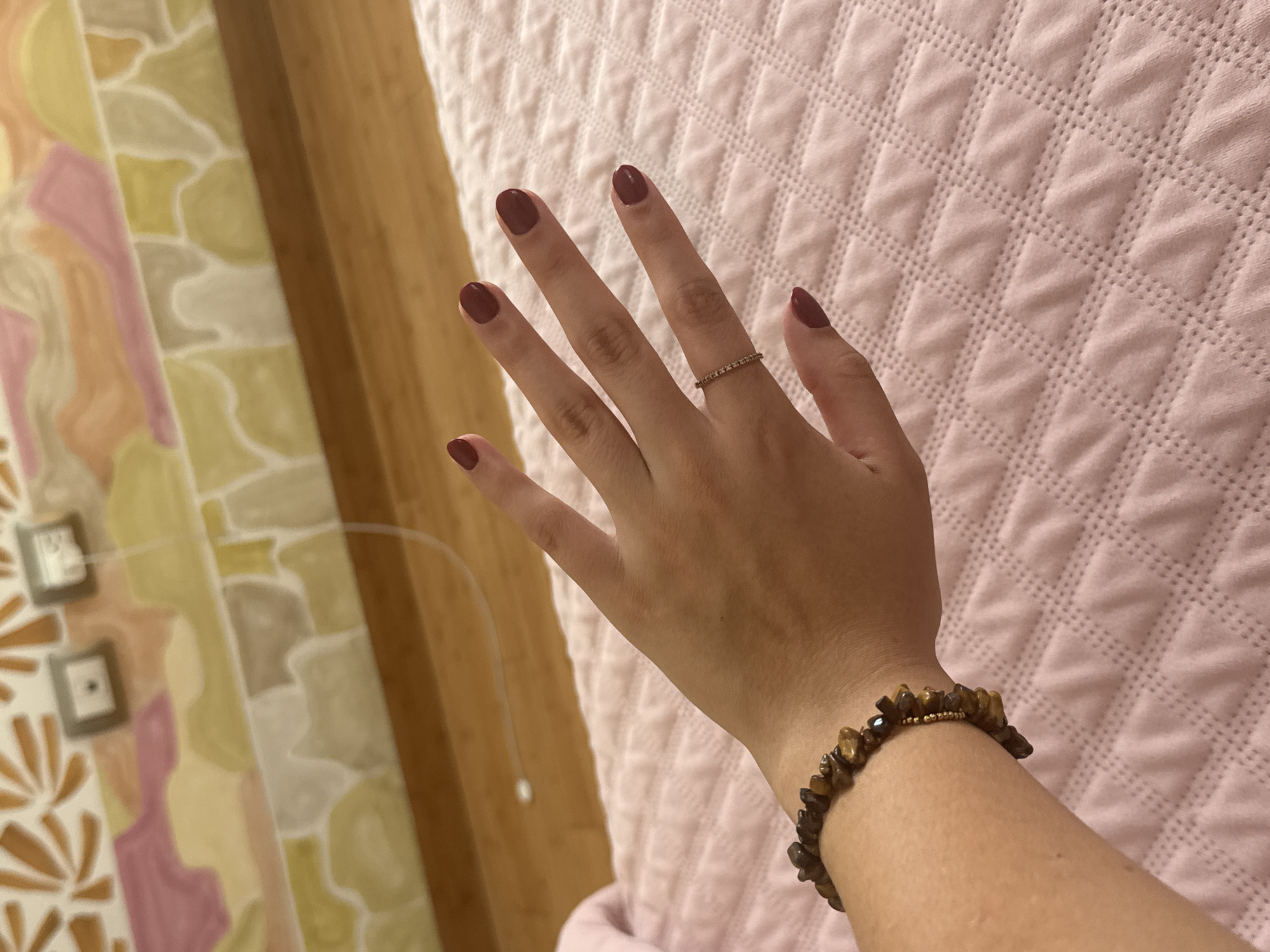 A person's hand and arm are positioned against a pink textured bedspread. The hand has a ring on the ring finger and the fingernails are painted a dark red color. A bracelet with brown and gold beads is on the wrist. The background includes a patterned wall or curtain with shades of yellow, pink, and white. The location is San Bernardino Tlaxcalancingo, Mexico, based on the prompt.