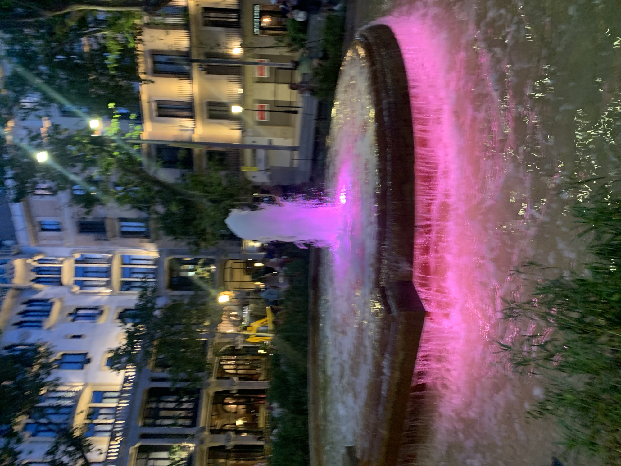 The image shows a fountain with pink lighting at night. The fountain is in an urban setting, with buildings visible in the background. The buildings are multi-story and appear to be apartments or businesses. There are trees and foliage surrounding the fountain, suggesting a park or plaza. People can be seen in the background, some sitting at tables, indicating an outdoor seating area. The overall atmosphere is festive and lively, with the bright pink lights adding a vibrant touch to the scene. The time of day is clearly night due to the lighting and the darkness of the sky. There is no visible text in the image.