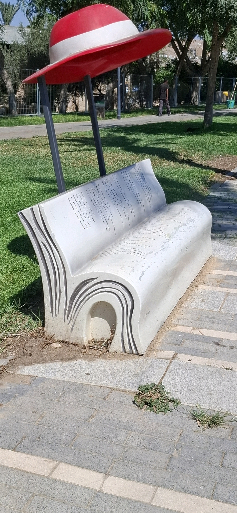 An artistic bench shaped like an open book is situated on a paved pathway in a park. The bench is white and has text inscribed on its surface, resembling pages. Behind the bench, a red and white hat-shaped sculpture is mounted on dark poles. The park features green grass, trees, and fencing. In the background, a man wearing dark clothing walks away from the camera. The scene appears to be in Dimona, Israel, as evidenced by the Hebrew text on the bench.