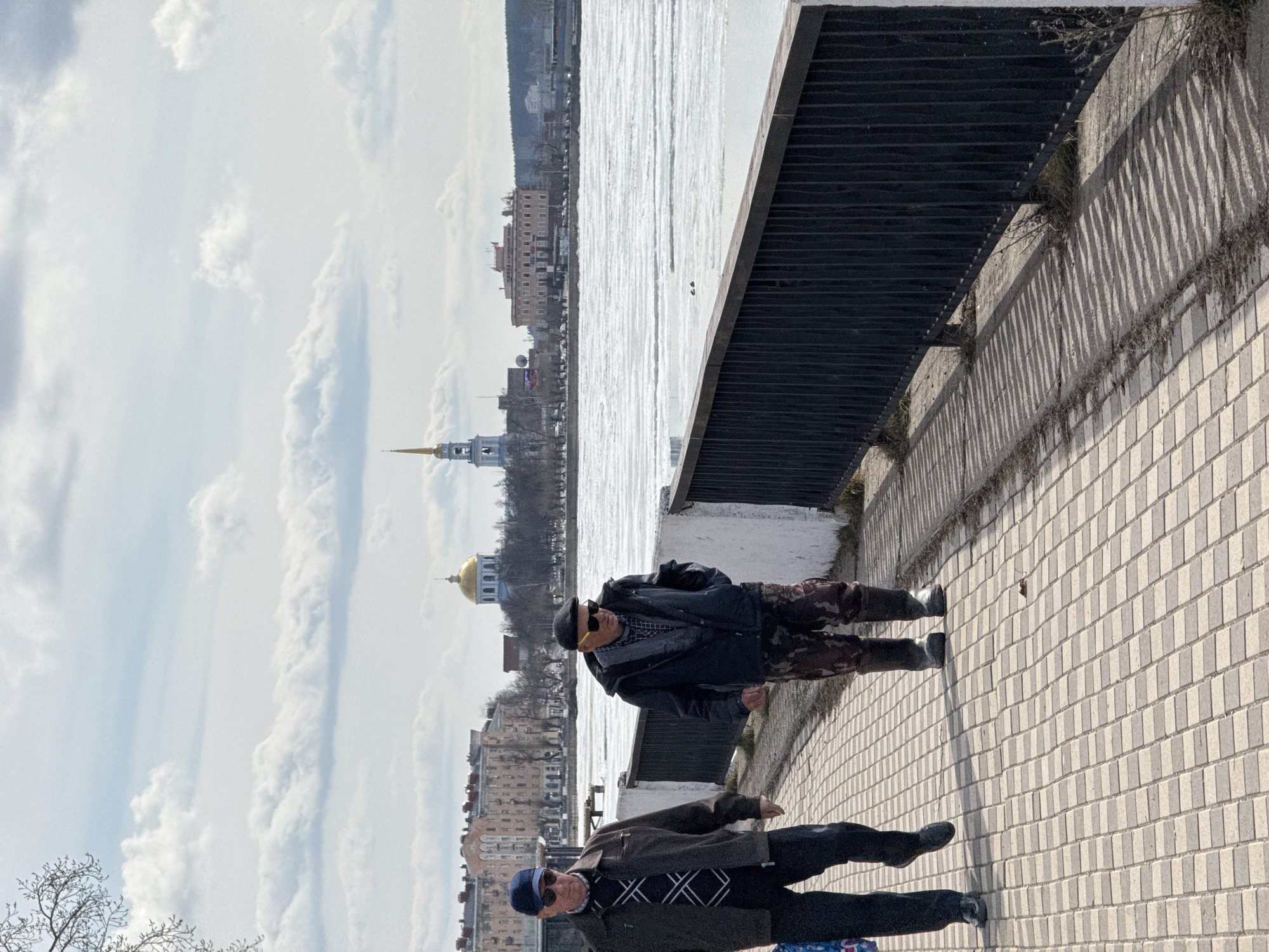 Two elderly men are walking along a paved walkway in Votkinsk, Russia, beside what appears to be the frozen Votkinsk Pond. The man closest to the camera wears a blue hat, sunglasses, a dark patterned sweater, a jacket, dark pants, and shoes. The other man wears a black hat, sunglasses, a dark jacket, camouflage pants, and shoes. They are walking beside a black metal railing, and in the distance, buildings and churches are visible. The sky is partly cloudy.