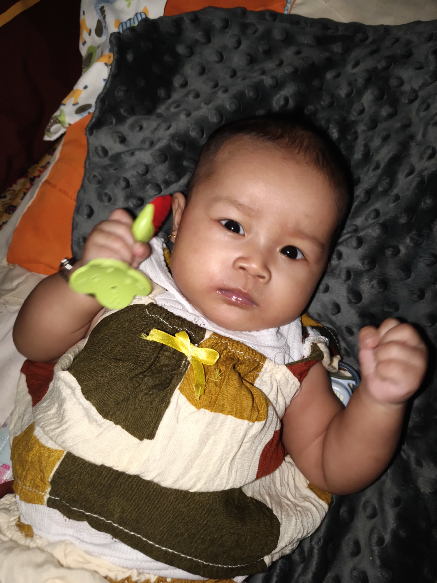 An infant lies on a dark, textured blanket in Barengkrajan, Indonesia, holding a green and red teething toy. The infant is wearing a dress with a patchwork design of off-white, mustard yellow, and dark olive green squares, accented with a small yellow bow. The background includes glimpses of pillows and blankets in various colors and patterns.