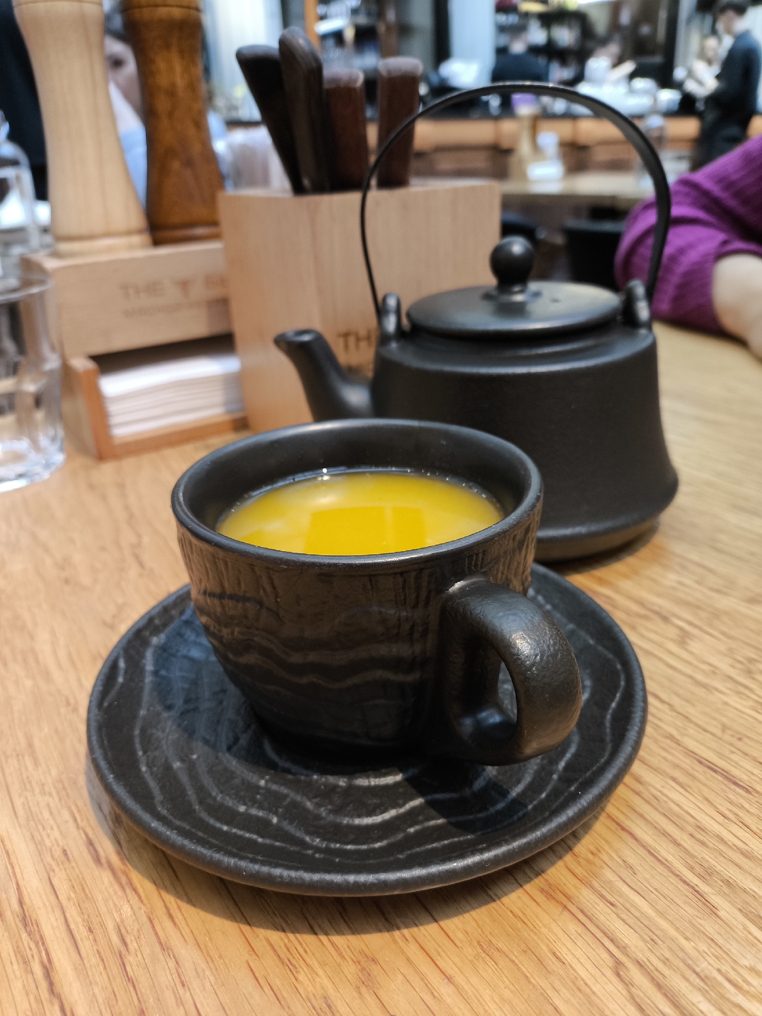 The image depicts a black ceramic tea cup and saucer, filled with yellow tea, alongside a black teapot on a wooden table. In the background, there's a wooden container holding utensils, a glass of water, and blurred figures of people, suggesting a restaurant setting. The specific location is unknown as the city is listed as Unknown, Unknown.