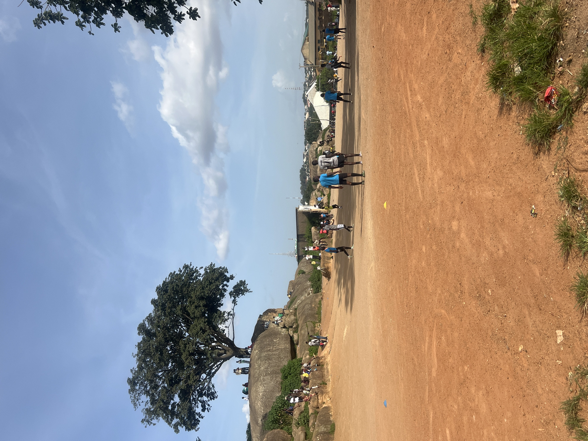 The image depicts a daytime scene in the city of Jos, Nigeria, characterized by a reddish-brown dirt field in the foreground, likely a makeshift sports ground. Several individuals, mostly young men and boys, are scattered across the field. Some are engaged in what appears to be a game of football, with a few players in motion, while others stand and observe or socialize.
In the midground, a significant rock formation dominates the landscape, topped by a large, mature tree providing shade. Numerous people are visible on and around these rocks, some seated and others standing. The rocks themselves appear weathered and are a prominent natural feature of the environment.
Further in the background, the urban environment of Jos is visible, with a cluster of buildings, some with corrugated iron roofs, and a distant transmission tower. The sky above is blue with scattered white clouds, indicating fair weather. The overall atmosphere suggests a community gathering and recreational activity in an open, natural, and urban setting.