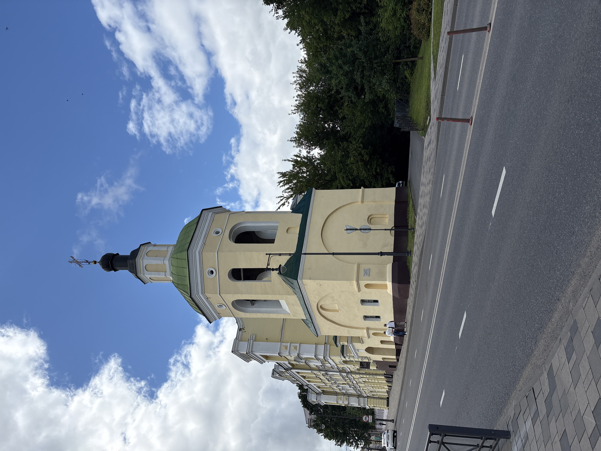 The image shows a light yellow church building with a green roof and a tall bell tower. The church is located in Lubnya, Russia, on a street with asphalt pavement and white lane markings. A sidewalk made of grey and black pavers runs alongside the street. Two people are walking on the sidewalk in front of the church. A white car is driving on the street in the distance. Lush green trees are visible behind the church. The sky is blue with scattered white clouds.