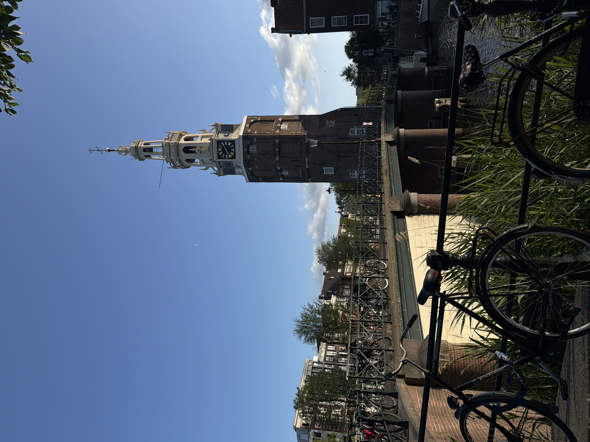 The image captures a view of the Montelbaanstoren, a historic tower located in Amsterdam, Netherlands. The tower, constructed of brick, features a clock face and a spire with a weather vane. It stands beside a canal, with a bridge in the foreground that spans the water. A row of bicycles is parked along the railing of the bridge, and some are also visible on the far side. The background reveals a line of traditional Dutch buildings and trees, bathed in the warm light of what appears to be late afternoon. The sky is clear blue with a few wispy clouds and a crescent moon visible. The overall scene conveys a sense of peaceful urban ambiance characteristic of Amsterdam.