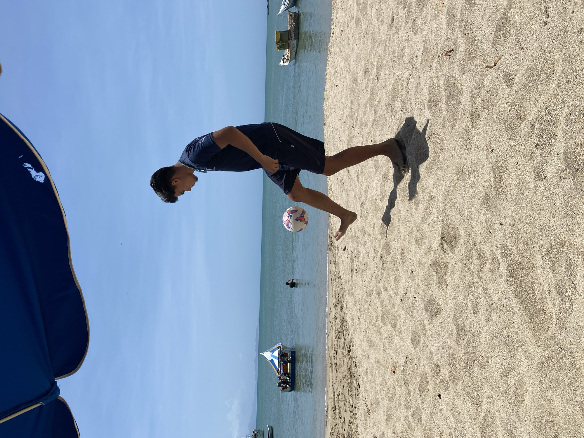 A teenager in a dark blue t-shirt and shorts is playing soccer on the beach in Juan Griego, Venezuela. The backdrop includes the blue ocean with several boats anchored, a person swimming in the water, and a clear, blue sky. The beach is sandy, with the shadow of the boy visible. Part of a blue umbrella is visible in the lower left corner.