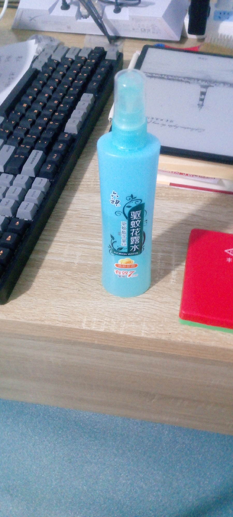 A light blue spray bottle is positioned on a wooden surface, to the right of a black keyboard. The bottle has Chinese characters and the words "FLORIDA WATER" printed on it. To the right of the bottle, a red object with a green edge is visible. In the background, a tablet device displays an illustration of a lighthouse. The scene appears to be an indoor desk area.