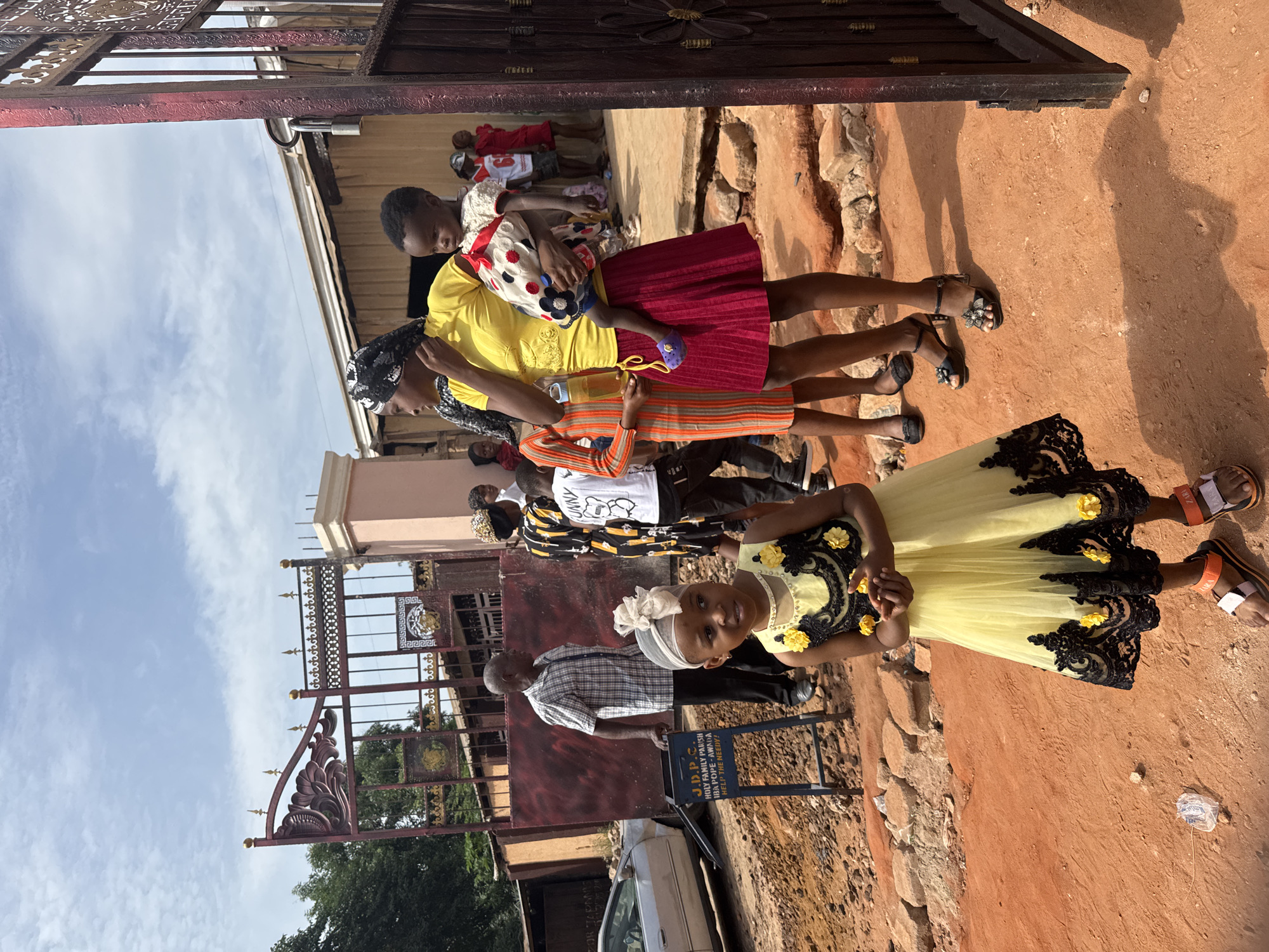 The image shows a group of people in Nkpor, Nigeria, standing outside a building with a brown gate. The gate has decorative metalwork. A young girl in a yellow dress with black lace and yellow flower accents stands in the foreground, looking at the camera. Behind her, an older man in a checkered shirt stands next to a blue sign. Further back, there are several other people, including women and children. One woman is wearing a yellow shirt and red skirt, carrying a child with a polka-dot bag. The ground is reddish-brown. A car is parked near the gate. The sky is visible in the background with scattered clouds.