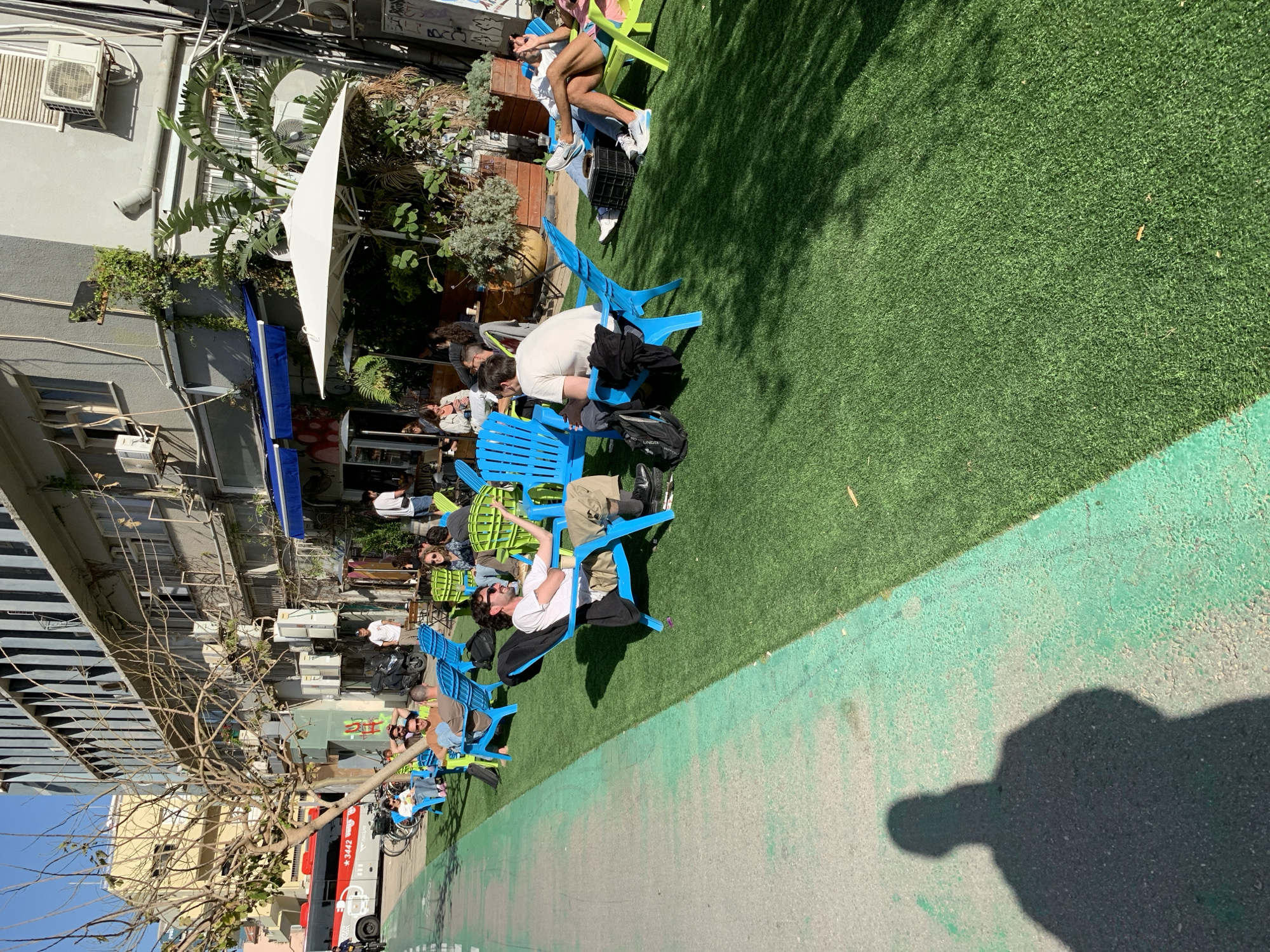 The image captures a street scene in Tel Aviv, Israel, featuring a line of people sitting in bright blue plastic chairs along a section of artificial grass laid adjacent to a sidewalk. Patrons are sitting, with a mix of clothing styles and postures. Some are interacting with each other. The background is composed of building facades, typical of Tel Aviv architecture. A large white umbrella provides shade over some of the seating, while the building walls are dotted with air conditioning units. A red bus is visible near the bottom of the frame. A shadow of a person is cast in the bottom right corner.