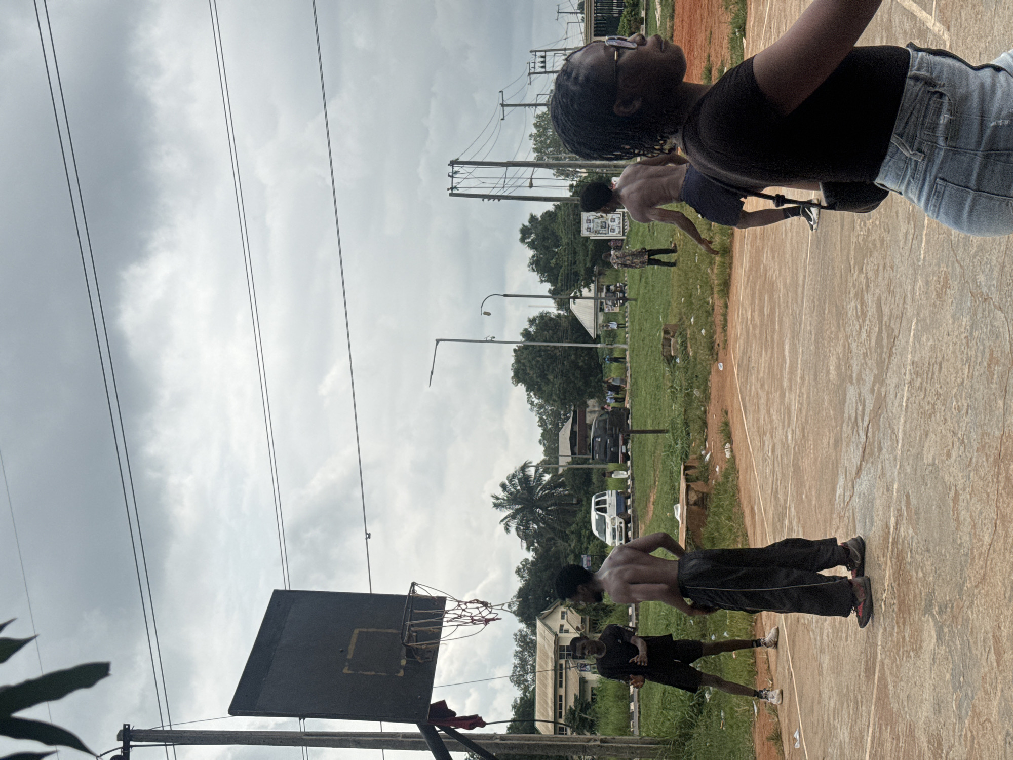 In Amansee, Nigeria, on a cloudy day, a group of people are gathered on an outdoor basketball court. In the foreground, a Black woman with braided hair and glasses is looking to her right, her shoulder and upper torso visible. Behind her, several Black men are engaged in a basketball game. One man, wearing black shorts and grey pants, is looking towards the basket. Another man, shirtless and wearing black shorts, is facing away from the camera. Further back, another man in a patterned shirt stands on the court. The basketball court is made of concrete and is surrounded by a grassy area. In the background, there are buildings, trees, and utility poles with power lines. Two vehicles, a white van and a dark SUV, are parked on a road in the distance, and a few other people are visible in the background. The overall scene depicts a casual outdoor activity in a semi-urban environment.