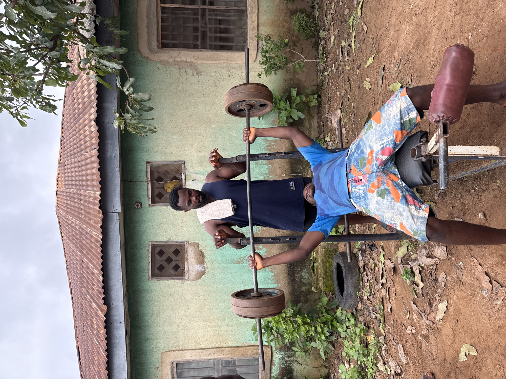 Two adult males are engaged in weightlifting outdoors in Akwanga, Nigeria. The man on the bench is performing a bench press with a barbell, while the man standing behind him is spotting him. Both individuals are Black. The background consists of a light green, weathered building with a rusty corrugated metal roof and two windows. The ground is composed of reddish-brown soil with scattered dry leaves. Lush green vegetation is visible on the left and right sides of the scene. A used tire lies on the ground near the bench.