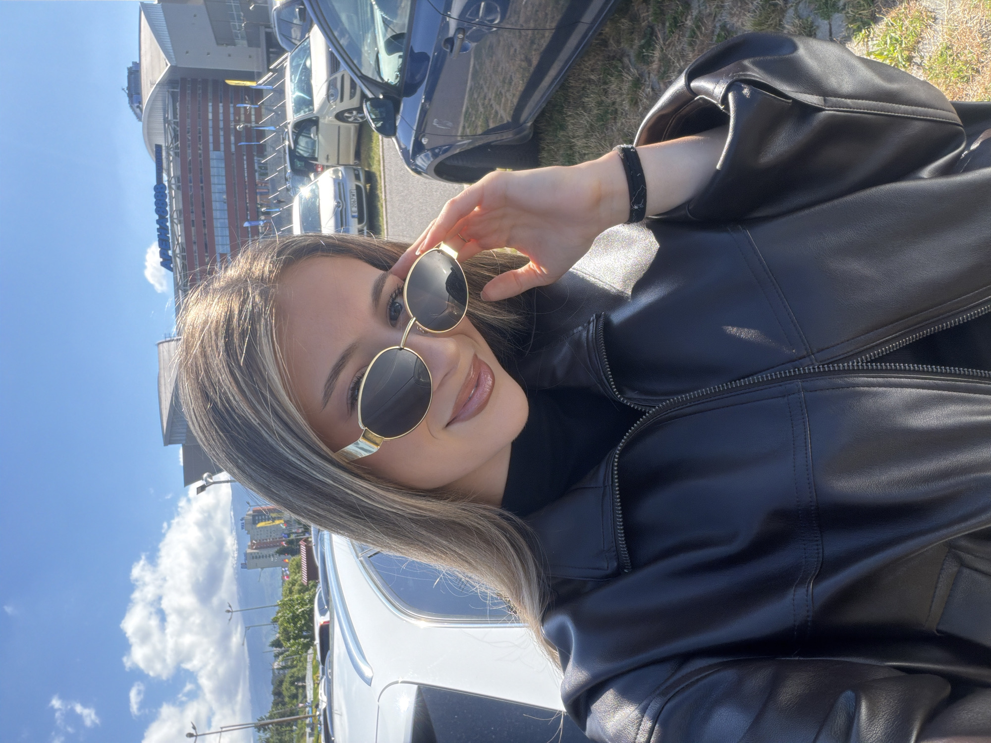 The image is a selfie of a woman in front of the Arena Armeec in Sofia, Bulgaria. She is wearing a black leather jacket and gold-rimmed sunglasses, which she is holding with her right hand. She has long, light brown hair with blonde highlights. Behind her are cars and the Arena Armeec, a large indoor arena with a glass facade. The sky is blue with a few clouds.