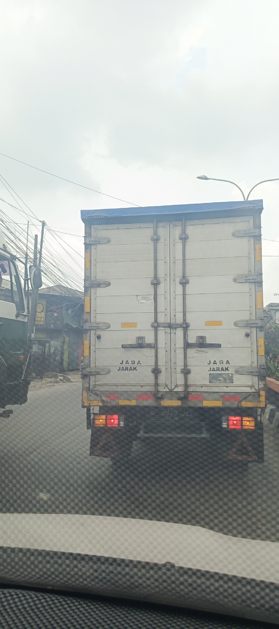 The image depicts the rear view of a white box truck on a city street in Sukaresmi, Indonesia. The truck has a blue tarp covering its roof. Text on the back of the truck reads "JAGA JARAK" in two locations, which translates to "KEEP DISTANCE" in English. The truck's license plate is partially visible and appears to read "B 9975 JZ". Red and yellow reflective tape is present on the rear of the truck. In the background, there are overhead power lines and buildings, suggesting an urban environment. A green truck is partially visible to the left of the box truck. The sky is overcast.