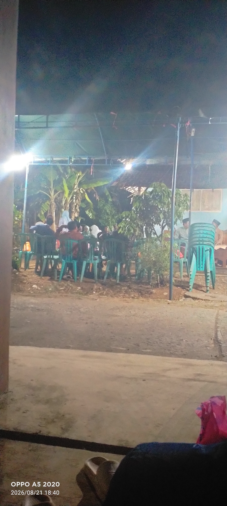 In Sowan Lor, Indonesia, a group of individuals are gathered outdoors, seated on teal plastic chairs under a covered canopy. Banana trees and other foliage are visible in the background, suggesting a tropical or subtropical environment. The scene is lit by artificial lights, creating a somewhat dim atmosphere. The composition implies a social gathering or event taking place at dusk or night. The timestamp at the bottom left corner of the image indicates it was taken on August 21, 2026, at 18:40.
