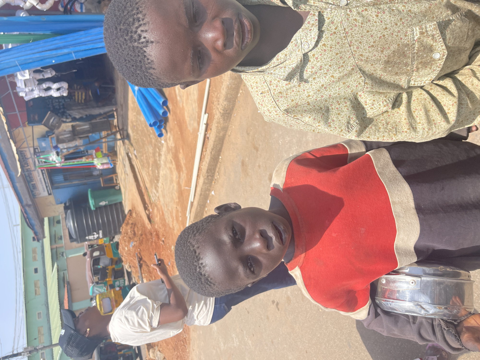 In Jos, Nigeria, on a dusty street, two young boys stand facing the camera. The boy on the left, with a red and black striped sweatshirt, holds a metallic container. The boy on the right wears a light green, floral-patterned shirt. Behind them, a woman in a white t-shirt and dark jeans looks away from the camera, holding a phone. The background reveals a bustling street scene with a blue building adorned with signs and a blue awning. Various objects are visible, including stacks of blue pipes, a black water tank, a green trash can, and a yellow auto-rickshaw. The ground is covered in dirt and gravel, characteristic of an outdoor urban environment.