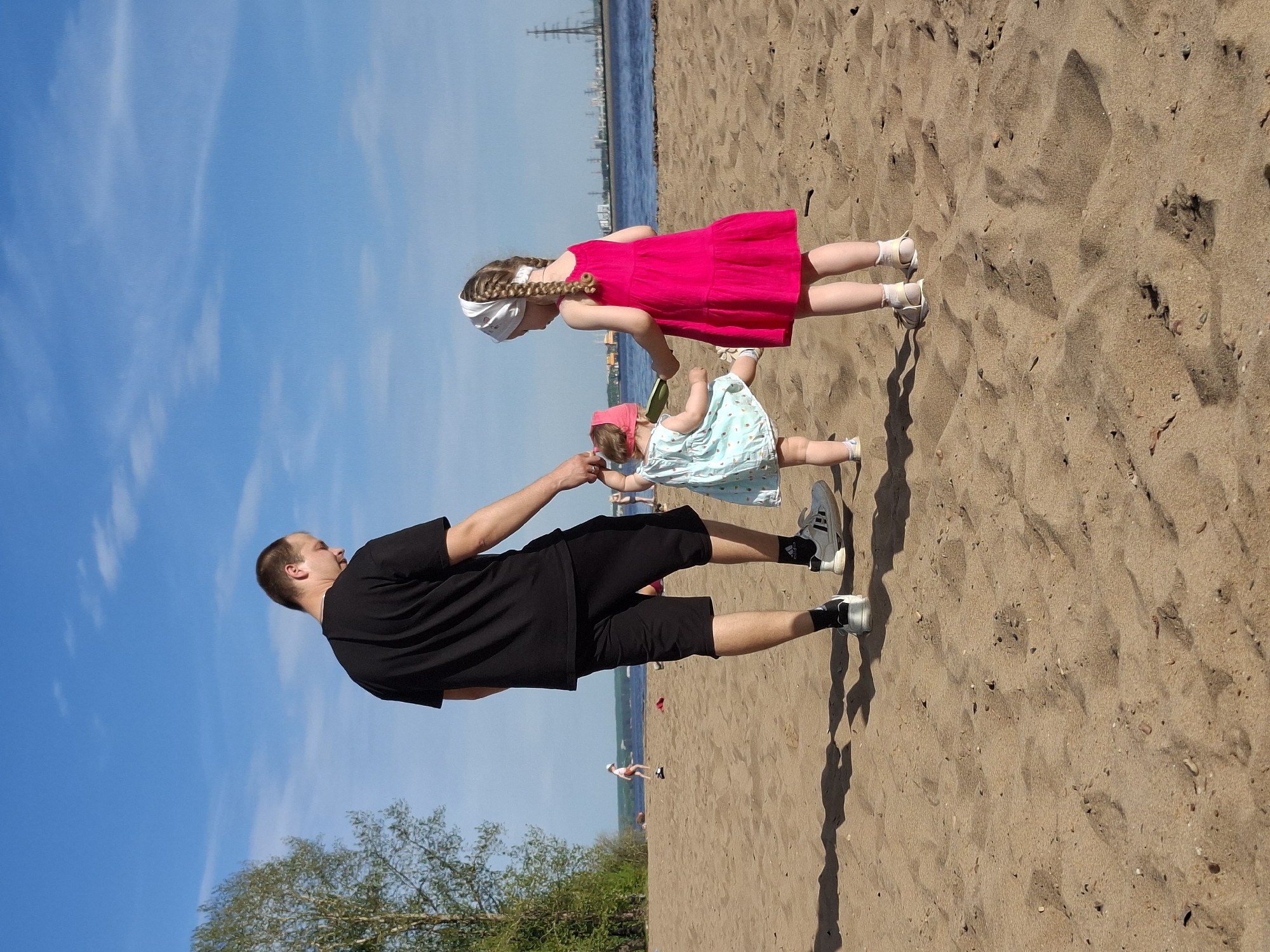 The image depicts a beach scene in Noviy, Russia, with a man and two young girls interacting on the sand. The man is wearing a black t-shirt, black shorts, and light-colored sneakers. He is standing and appears to be holding or assisting the younger of the two girls. The younger girl is wearing a light-colored dress with a pink hat. The older girl is wearing a pink dress and a white headband. The background features a body of water and a coastline in the distance. The sky is blue with some clouds. The beach is sandy, and there are some trees visible on the left side of the frame.