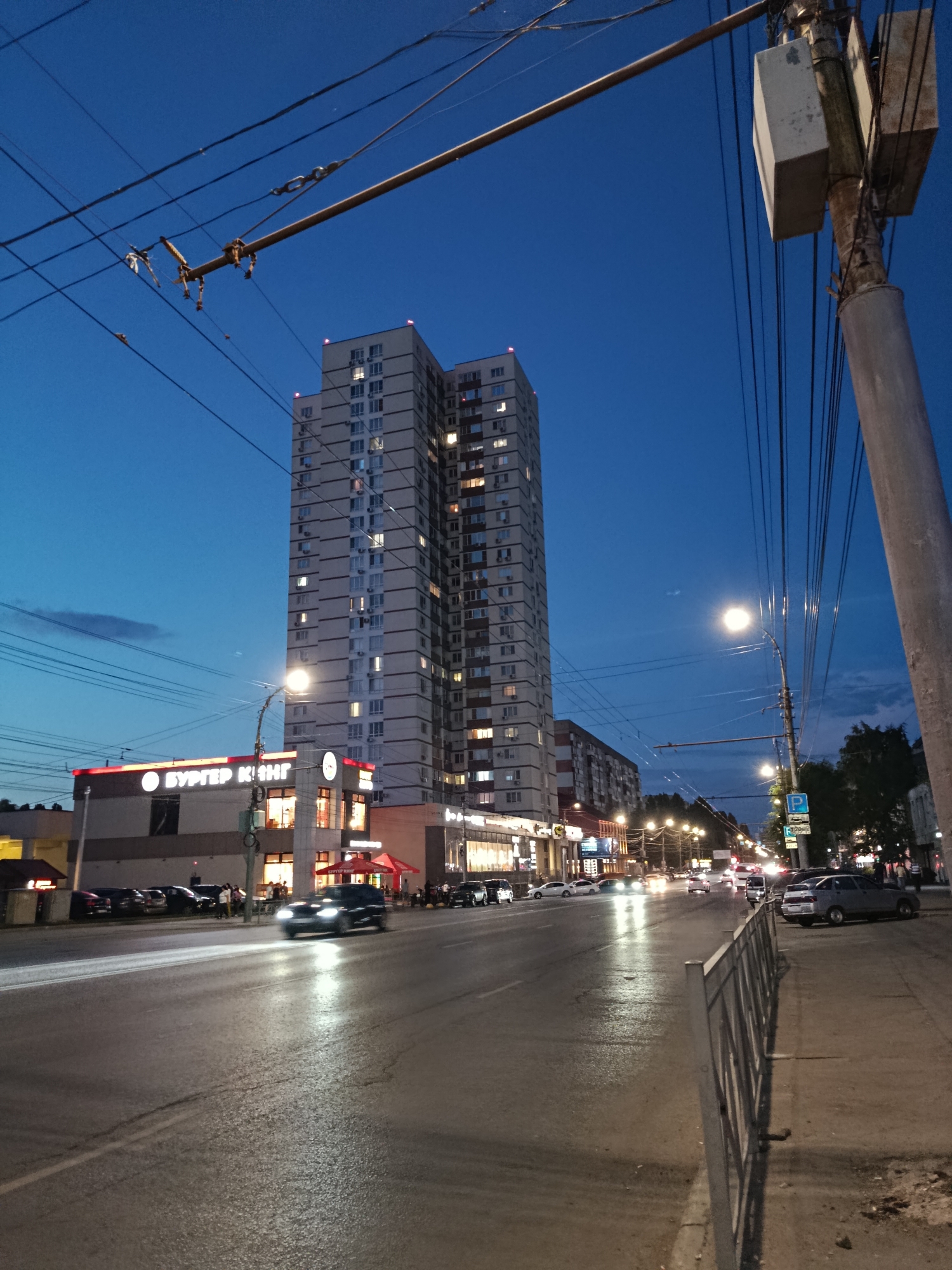 The image shows a wide street in Saratov, Russia, at dusk. The sky is a deep blue, with a few wispy clouds visible. Overhead, a complex network of trolleybus wires crisscrosses the frame, supported by poles.
On the left side of the street, a multi-story building with a Burger King restaurant on the ground floor is prominent. The Burger King sign is illuminated, and people can be seen inside the restaurant. Further down the street, other businesses with illuminated signs are visible. A tall, modern apartment building with many windows, some lit up, stands behind the Burger King establishment.
The street itself is wet, reflecting the headlights of passing cars. Several vehicles are on the road, including a black car in the foreground with its headlights on, creating a bright streak on the wet pavement. On the right side of the street, a sidewalk with a metal fence runs parallel to the road. Several cars are parked along the sidewalk. Streetlights illuminate the scene, casting a warm glow. Trees are visible in the distance on the right side of the image. The overall impression is that of a busy urban environment at the end of the day.