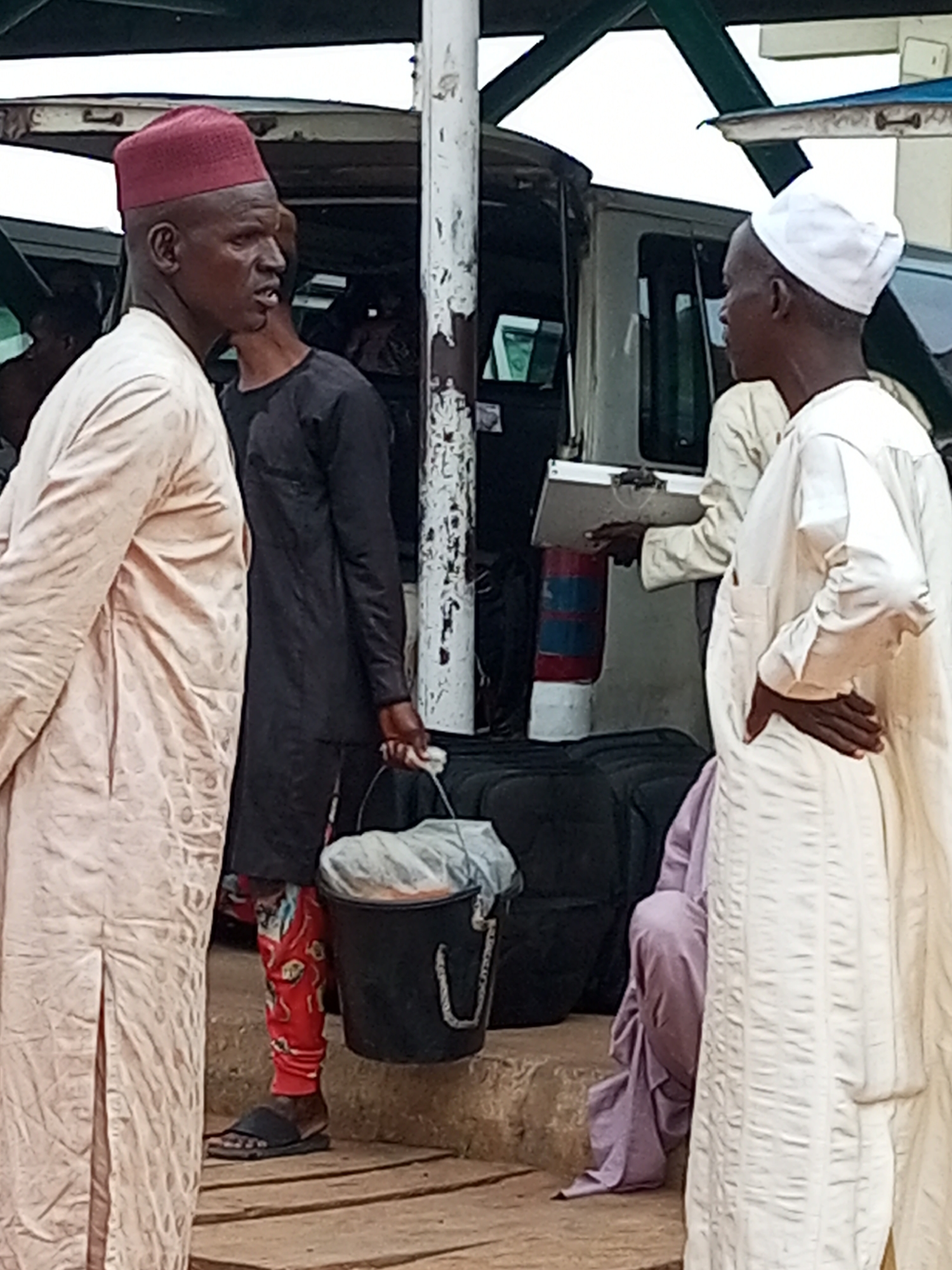 In Gombe, Nigeria, at what appears to be a transportation hub or border crossing, two men in traditional attire are captured in a photograph. The man on the left wears a maroon cap and a light-colored robe with subtle patterned embroidery. He is looking to his right, his mouth slightly open as if speaking. To his right, a man in a black garment stands behind him, also looking to the right. He is holding a black bucket with a white plastic bag inside, and his right hand is resting on the bucket's handle.
In the background, several vehicles are visible, including a white van with its doors open. Luggage, possibly bags and suitcases, is stacked near the vehicles. Another man, wearing a white cap and robe, stands in the middle ground, also looking to his right, with his right hand on his hip. Further to the right, a person in a purple garment is seated. The ground appears to be a mix of concrete and wooden planks. The overall scene suggests a moment of transit or waiting, common in a busy transportation area in Gombe.