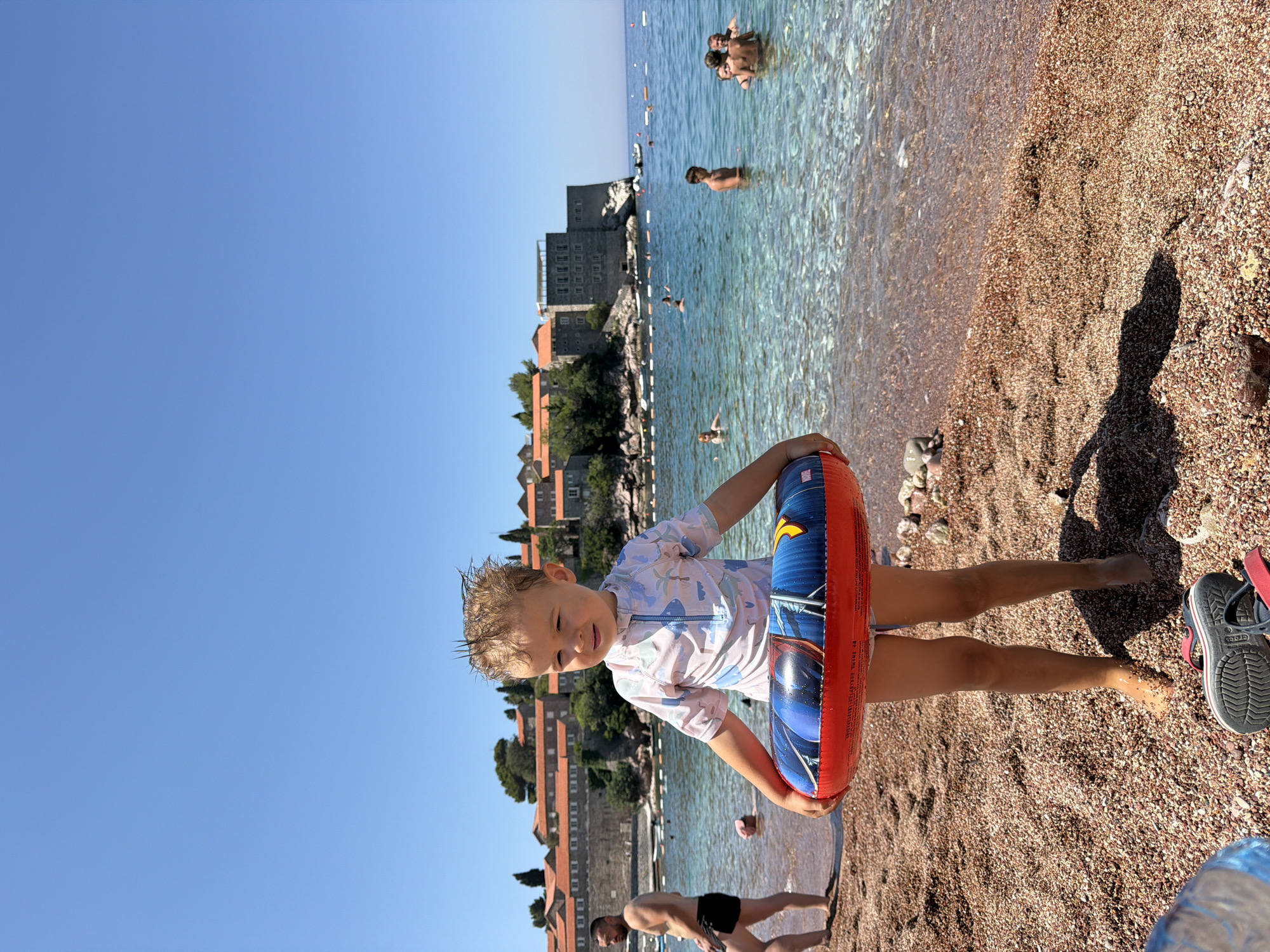 A young child stands on a pebble beach, holding a red and blue inflatable ring with a Marvel comic character design. The child is wearing a light-colored rashguard with a blue zipper and patterned print, and white shorts. Behind the child, the clear turquoise water of the Adriatic Sea is visible, with several people swimming and bathing. A man in black swim trunks walks along the shoreline to the left of the child. In the background, a historic island town with orange-tiled roofs and stone buildings is perched on a rocky outcrop, surrounded by the sea. The sky is a bright, clear blue. The overall scene depicts a sunny day at the beach in Rađenovići, Montenegro.