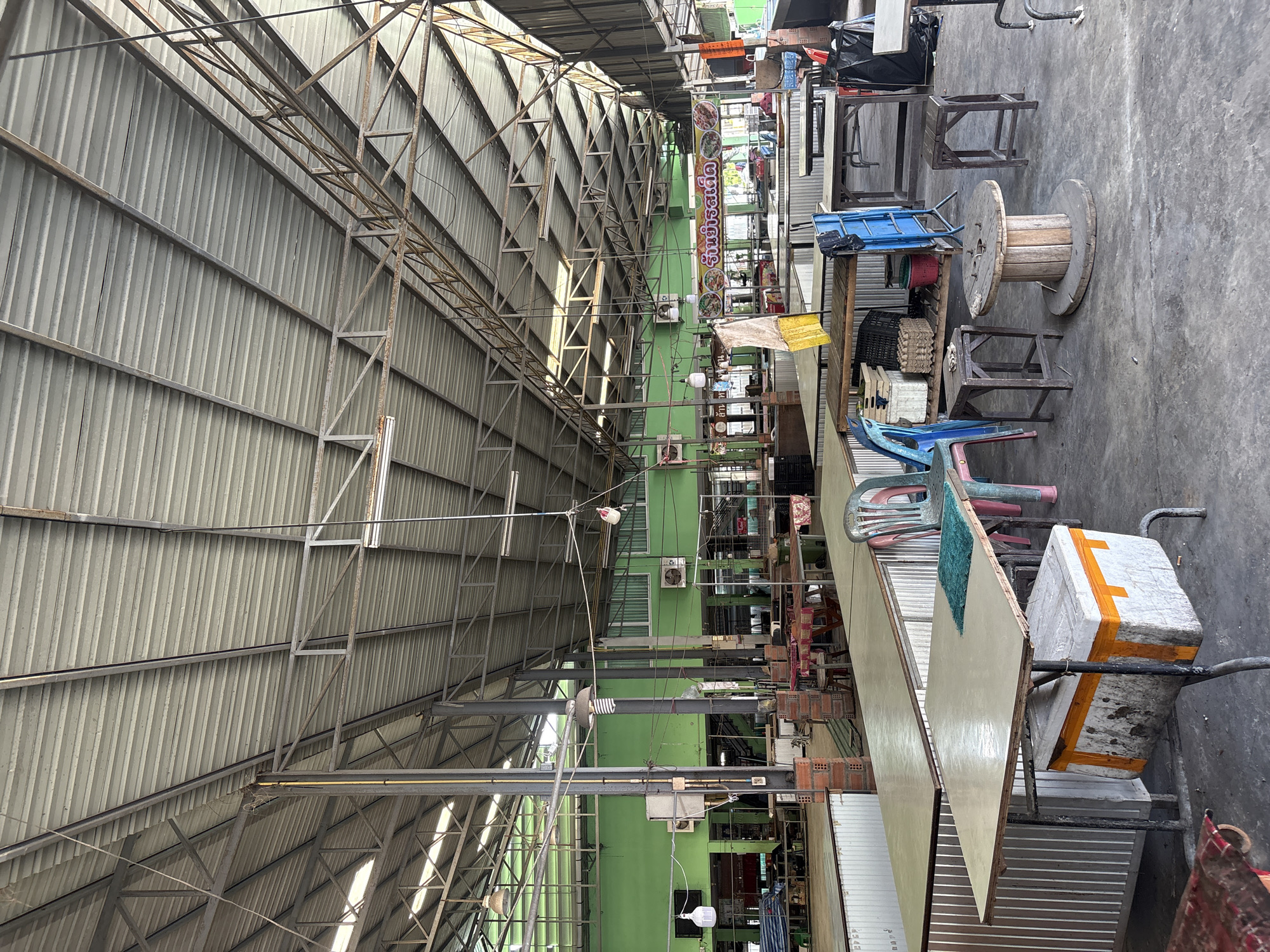 This is a wide shot of an empty outdoor market stall area in Chalong, Thailand. The market is covered by a corrugated metal roof supported by a metal frame structure. The background features a long, vibrant green building with multiple windows and air conditioning units. In the foreground and midground, numerous empty tables and chairs are set up, suggesting a retail or food service environment. Some tables have coolers or storage containers beneath them, and a few chairs are occupied by small stools. A large wooden spool is placed in the center of the frame, and there are various baskets, crates, and trash bags visible. A banner with Thai text and images of food hangs across the market in the midground. The overall impression is one of a quiet, unoccupied market during daylight hours.