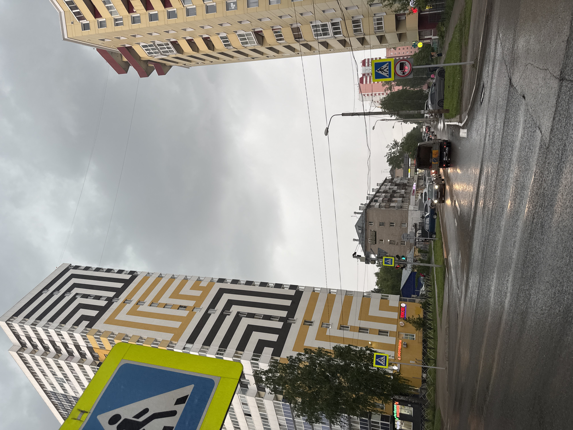 The image depicts a street scene in Perm, Russia, under an overcast, gray sky, likely during the late afternoon or early evening following a rain shower, as evidenced by the wet asphalt road and illuminated vehicle headlights.
In the prominent left foreground, a modern, tall residential building stands out with a distinctive geometric facade featuring a complex pattern of black, white, and yellow lines. A blue pedestrian crossing sign on a yellow background is partially visible in the bottom left, positioned in front of this building.
The street, wet from recent rain, stretches into the distance, with several cars and a dark-colored van driving or stopped. Headlights of oncoming vehicles are clearly visible. Along the right side of the street, there's another pedestrian crossing sign, and a red circular sign indicating "No Buses" is posted near the sidewalk. A green traffic light is also visible further down the road.
Various other multi-story buildings line both sides of the street, including a light-colored building with red architectural elements in the upper right. Overhead wires and streetlights crisscross the sky, and some trees and green spaces are visible along the road.
On a yellow building in the middle distance, two signs with Cyrillic text are visible: "ПИВОН" (PIVON, likely a beer store or bar) and "ЗООТОВАРЫ" (ZOOTOVARY, meaning pet supplies).