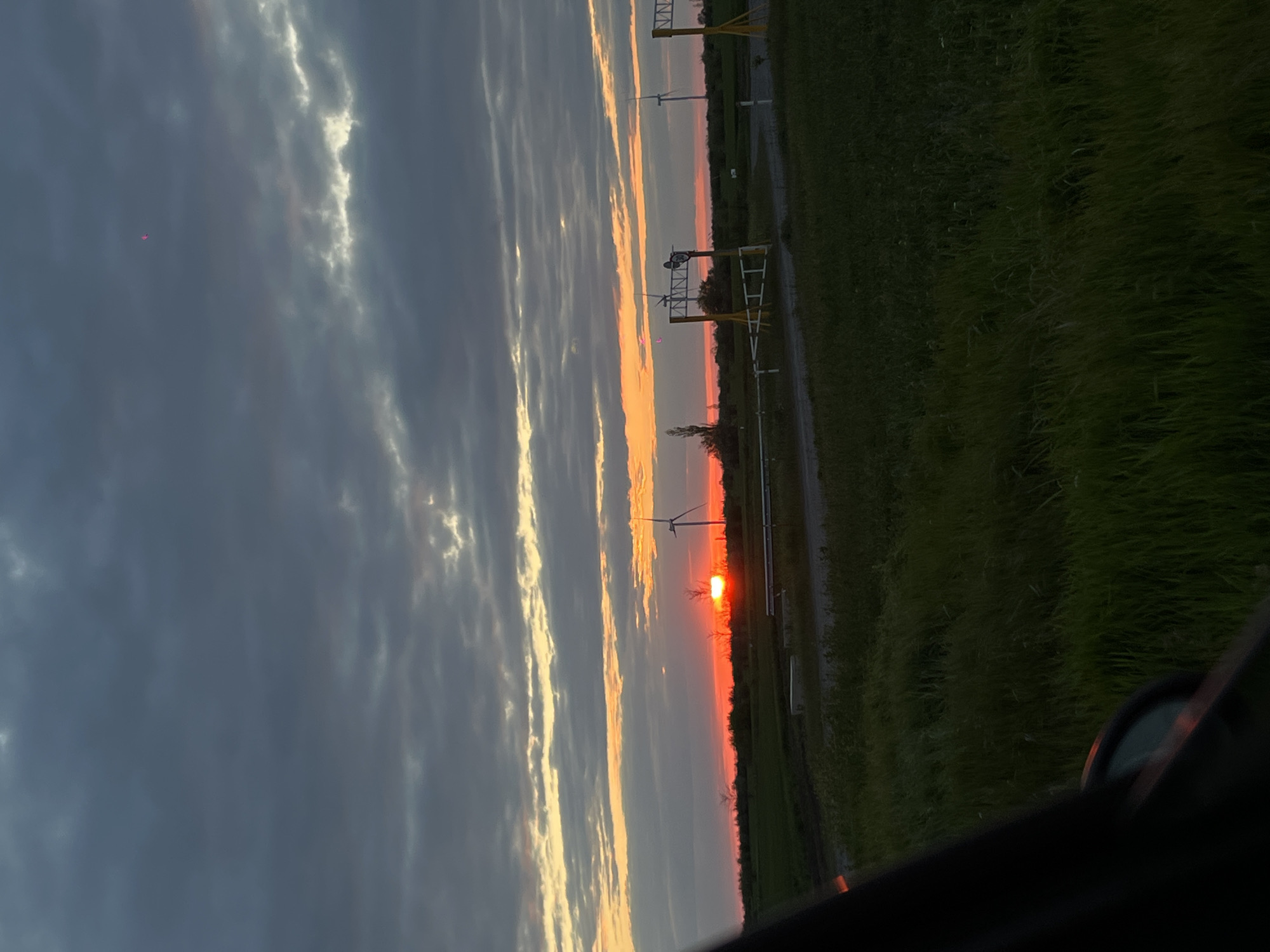 The image depicts a sunset over a field in what may be the outskirts of a city. The sky is a mix of dark clouds and orange-yellow light, indicating the setting sun. Wind turbines are visible in the distance, suggesting a rural area. There is also a metal structure similar to electrical infrastructure along with grass on the right side. A reflection can be seen, suggesting the photo was taken from inside a car. The location is unknown.