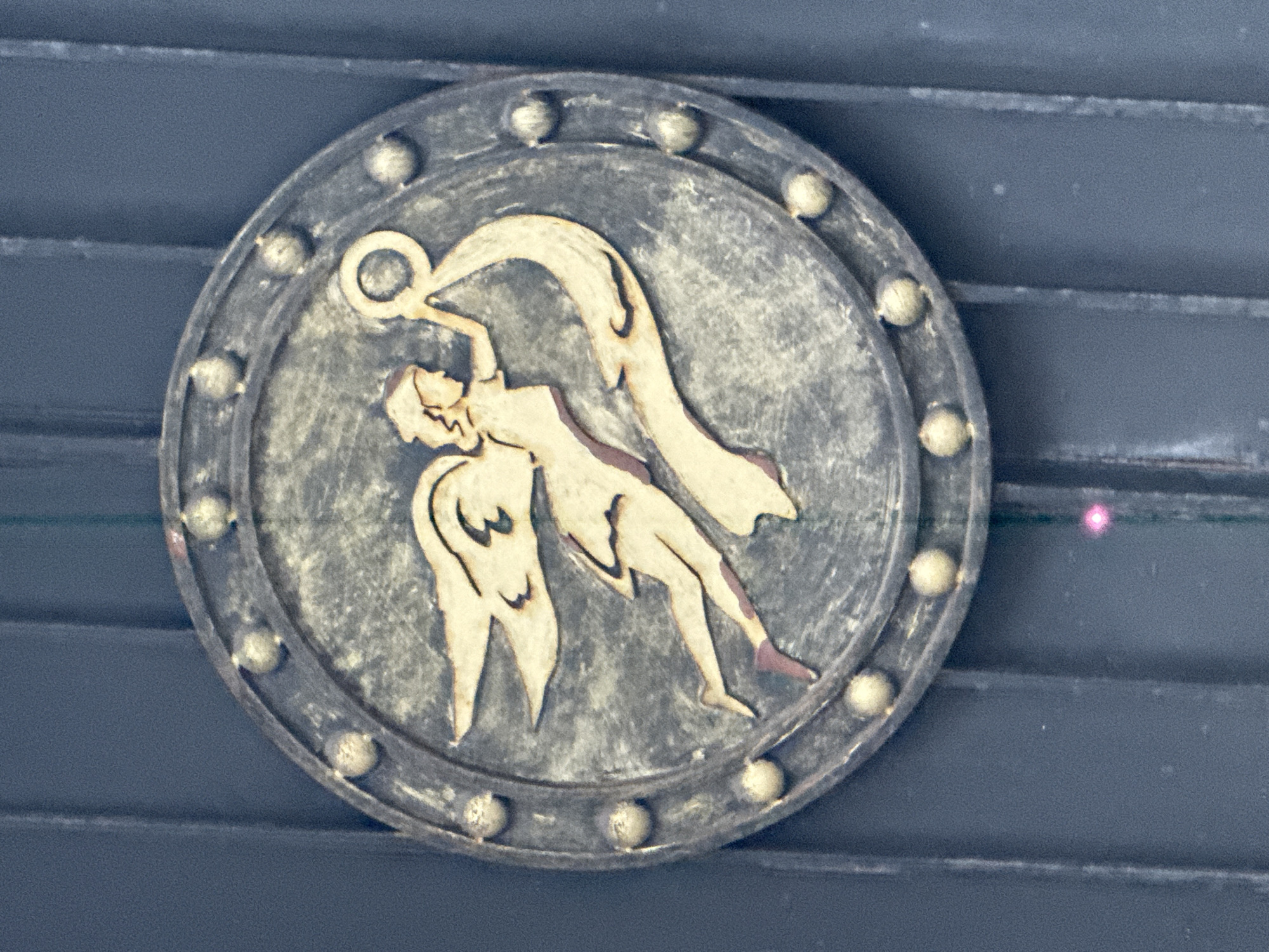 A circular, antique-styled medallion is centered against a dark, horizontally grooved background, likely wood or a similar architectural surface. The medallion appears to be crafted from dark gray, weathered metal, exhibiting a textured and aged patina. Its raised outer rim is adorned with evenly spaced, small, spherical studs, which also suggest a metallic material.
Within the medallion, a stylized winged figure is depicted in a light beige or off-white color, accented with dark outlines and subtle darker shading on areas such as the lower legs and feet. The figure possesses a human or humanoid form, possibly representing an angel or a spirit, and is captured in a dynamic, upward-reaching posture. One arm is extended upwards, grasping a circular object, which could be a halo or a ring. The body is angled, and the wings are spread, conveying a sense of movement, ascent, or flight. The background inside the medallion matches the weathered dark gray texture of its frame.
No visible text is present in the image. The scene suggests a decorative or symbolic artwork mounted on a wall. The image was captured in Moscow, Russia.