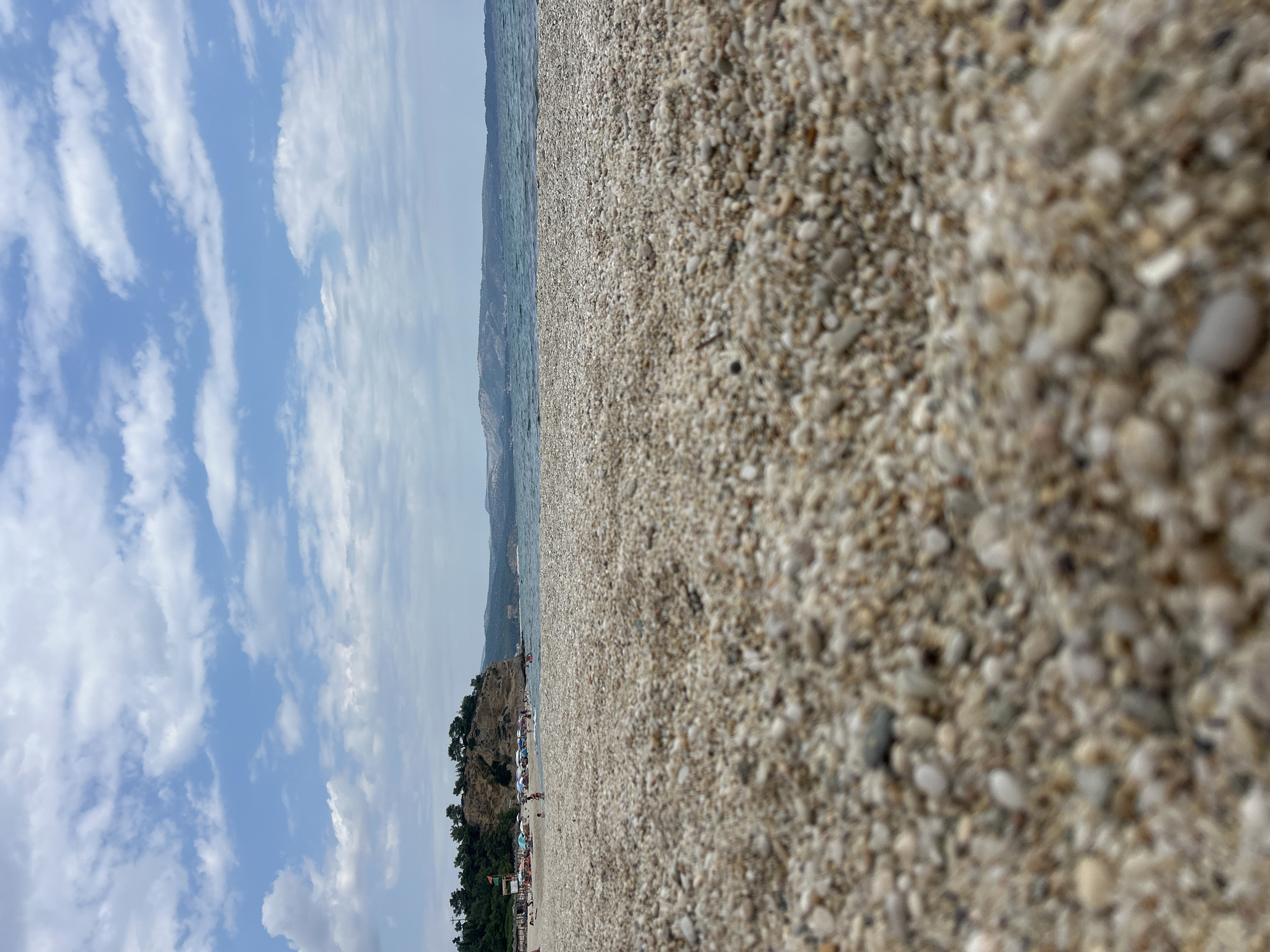 A low-angle, full outdoor shot captures a beach with a prominent focus on the coarse, pebble-filled sand in the foreground. In the middle ground, the sea stretches out under a partly cloudy sky, with a distant coastline visible. The background features a rocky cliff dotted with trees, and further back, a hazy mountain range. A small cluster of beach umbrellas and a few people are visible on the sand near the water's edge. The overall atmosphere is that of a calm summer day at a coastal location.