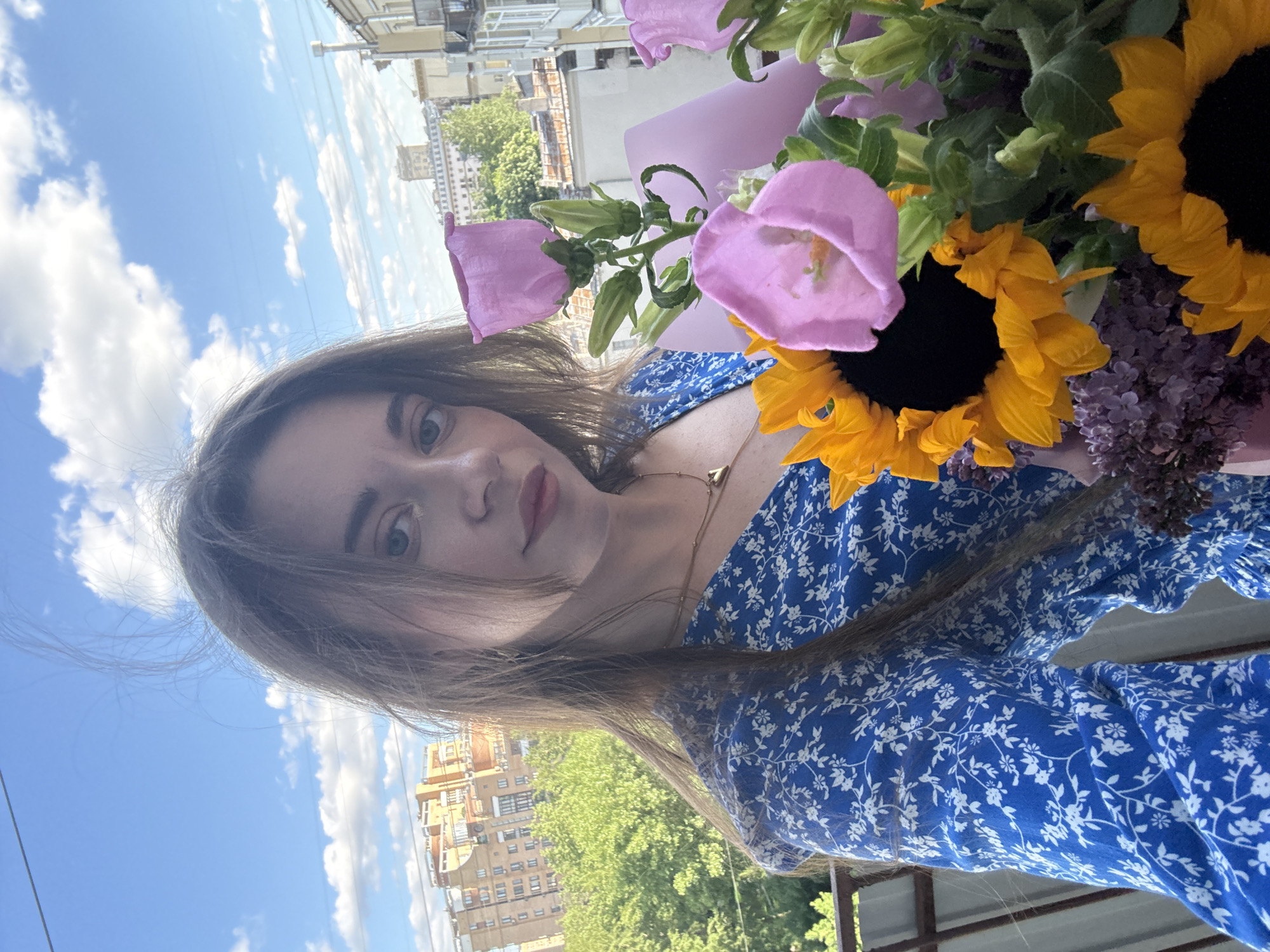 A woman is taking a selfie outdoors with a bouquet of flowers that includes sunflowers and purple flowers. The woman is wearing a blue and white floral dress. In the background, a city scene is visible under a blue sky with clouds. The location appears to be Moscow, Russia, based on the architectural style of the buildings.