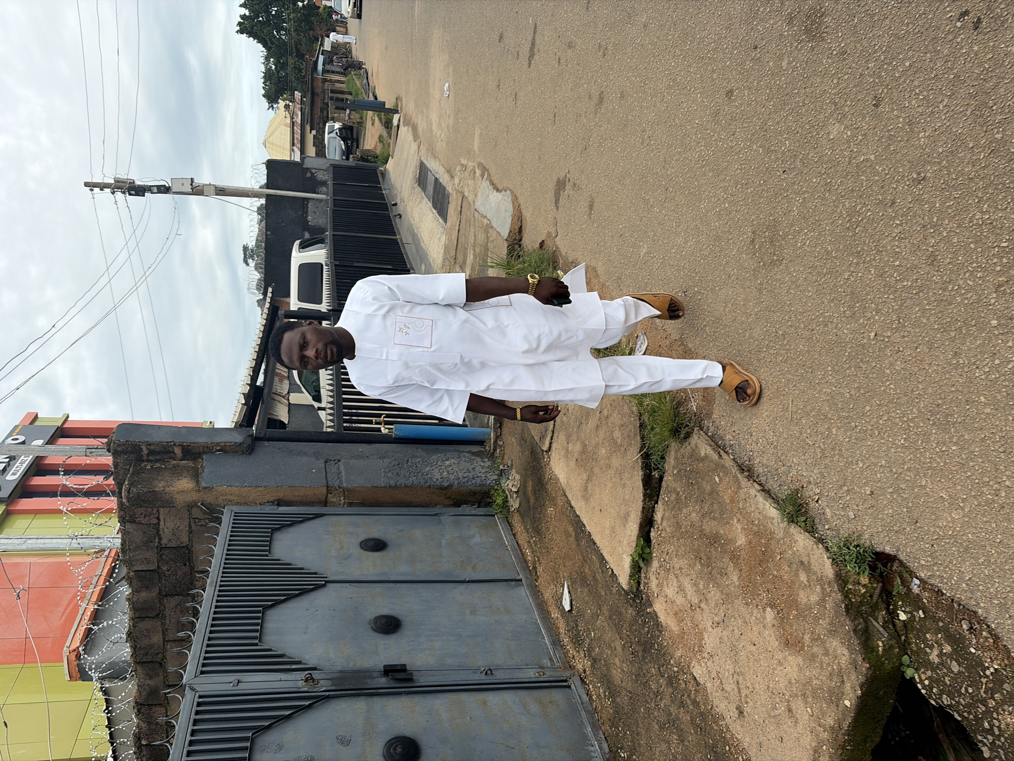 A full-body, eye-level shot shows a Black man walking on a paved street in the city of Jos, Nigeria. He is wearing a white traditional outfit with matching pants and a short-sleeved tunic that has a subtle embroidered design on the left breast pocket. He has brown sandals on his feet, and on his left wrist, he wears a gold watch. The man is looking forward, and his left hand is holding a dark-colored mobile phone. He is walking past a gray metal gate with pointed finials, which is part of a wall with barbed wire on top. Behind the wall is a building with orange and yellow facades. The street is lined with other buildings and parked vehicles. The sky is overcast.