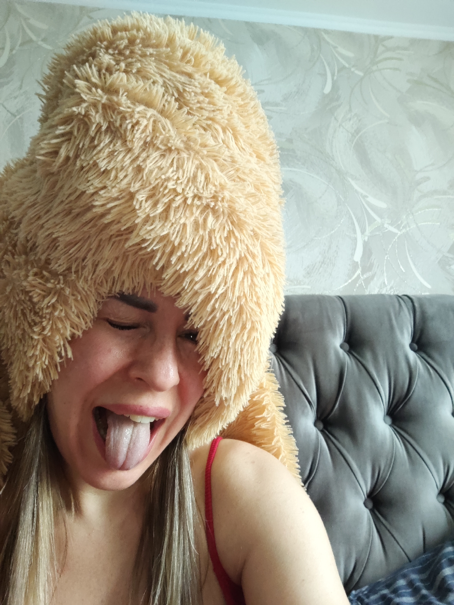 The image is a close-up, medium shot of a young woman wearing a fluffy, tan, novelty hat that resembles an animal's head. She is indoors, sitting against a tufted grey headboard, likely in a bedroom. The wall behind her has a light-colored, patterned wallpaper. The woman is making a playful face, with her eyes closed and her tongue sticking out. She is wearing a red strap, possibly from a tank top or bra. Her blonde hair is visible, falling down past her shoulders. The lighting suggests it is daytime, and there is no indication of weather conditions. The overall mood is lighthearted and fun. There is no visible text in the image.