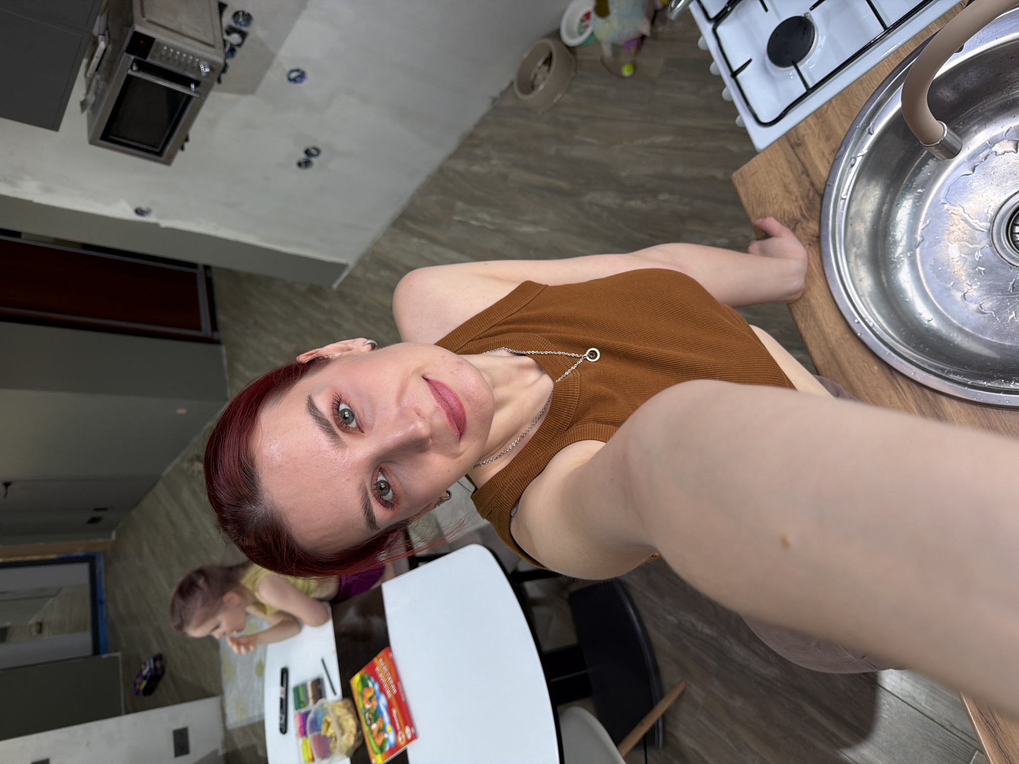 This selfie image captures a woman and a child in what appears to be a domestic kitchen and dining area in Chaikovsky, Russia. The woman, who is the primary subject, takes up the left and central portion of the frame. She has red hair, light eyes, and is looking directly at the camera with a subtle, pleasant expression. She wears a brown ribbed tank top and a silver chain necklace with a circular pendant. Her arm is extended as she holds the camera, with a small mole visible on her arm in one of the close-up crops.
In the background, a child with light hair, dressed in a yellow top, is seated at a white table, engrossed in an activity. Various art supplies, including crayons or paints in small compartments, and a book with colorful illustrations are scattered on the table. Visible Cyrillic text on the book cover includes "сказки" (tales).
The setting is a kitchen area, with a stainless steel sink, a flexible beige faucet, and a white gas stove with black grates visible to the right of the woman. The sink appears to have water droplets, suggesting recent use. A microwave oven is mounted above a counter in the upper left background. The countertops are a light wood pattern, and the flooring is a darker wood-patterned tile or laminate. Some walls have an unfinished or textured appearance, possibly indicating ongoing renovation. Recessed lights are visible on the ceiling. The overall lighting suggests it is daytime.