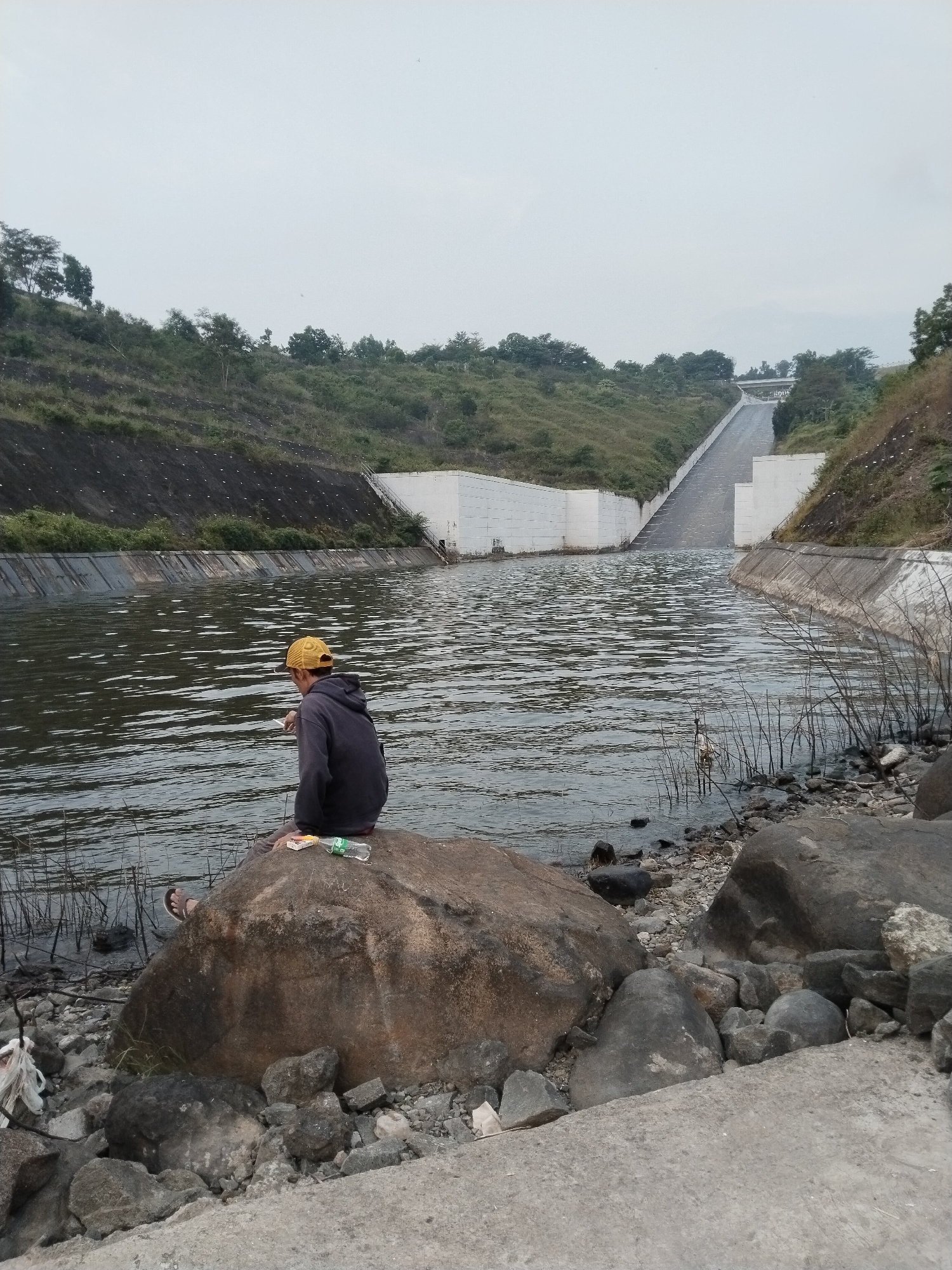 A person wearing a yellow cap and dark hoodie sits on a large rock at the edge of a canal, near Ganten, Indonesia. The canal is lined with concrete walls and slopes upward in the background, possibly part of a dam or water management system. The surrounding landscape includes grassy hills and trees. The foreground shows rocks and debris along the shoreline. A water bottle and a pack of cigarettes are visible on the rock.