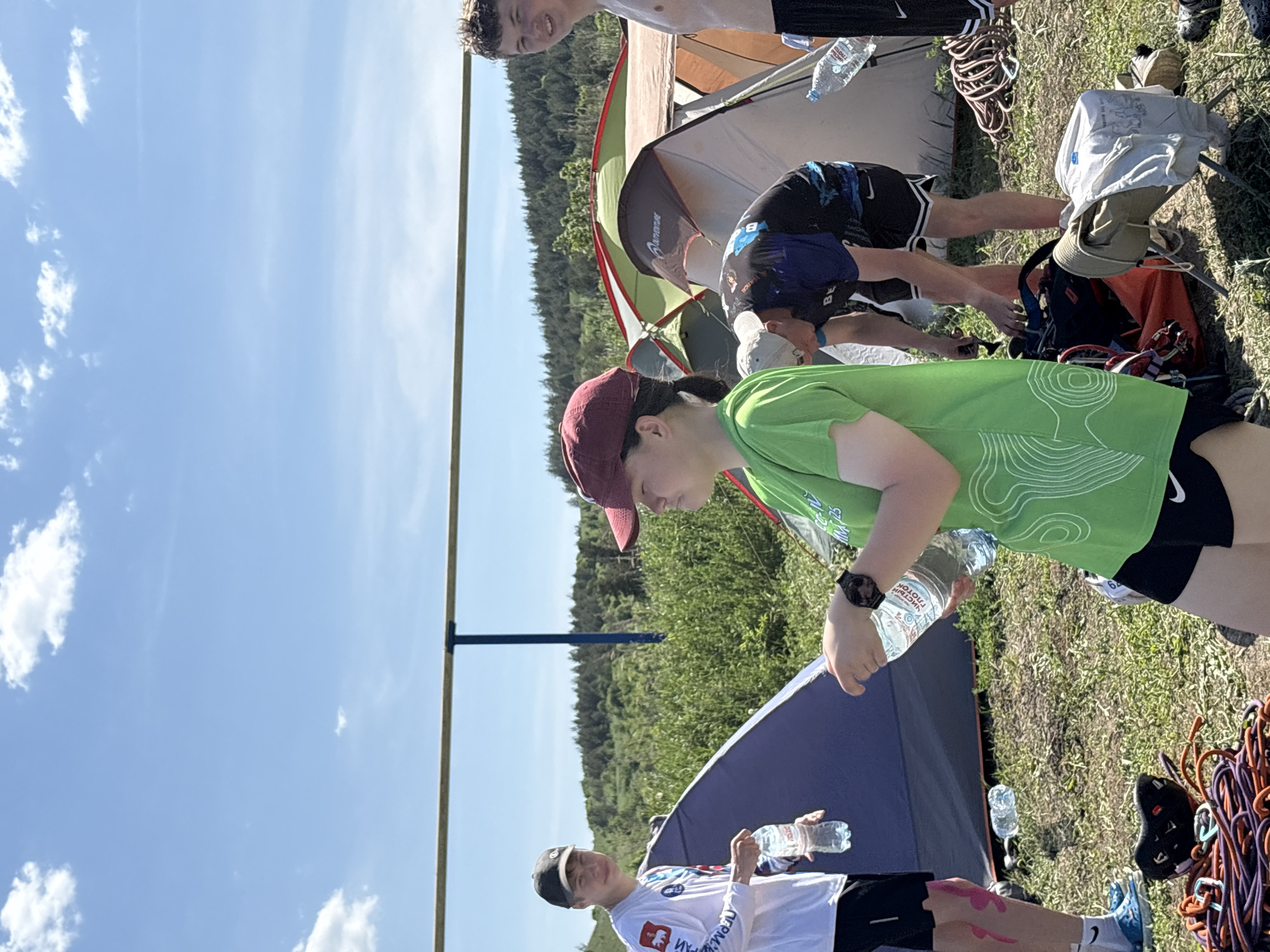 The image depicts an outdoor scene near what appears to be a camping area in Khvalynsk, Russia. Several people are visible. One person, wearing a green t-shirt, black shorts, and a red cap, is holding a water bottle. Another person, wearing a white jersey, black shorts, and a cap, is also holding a water bottle and standing near a blue tent. A third person is shirtless, while another is leaning into a tent. In the background, there is a green tent, trees, and a blue sky with clouds. A vertical pole is on the left side of the image. Climbing ropes and gear are lying on the ground.