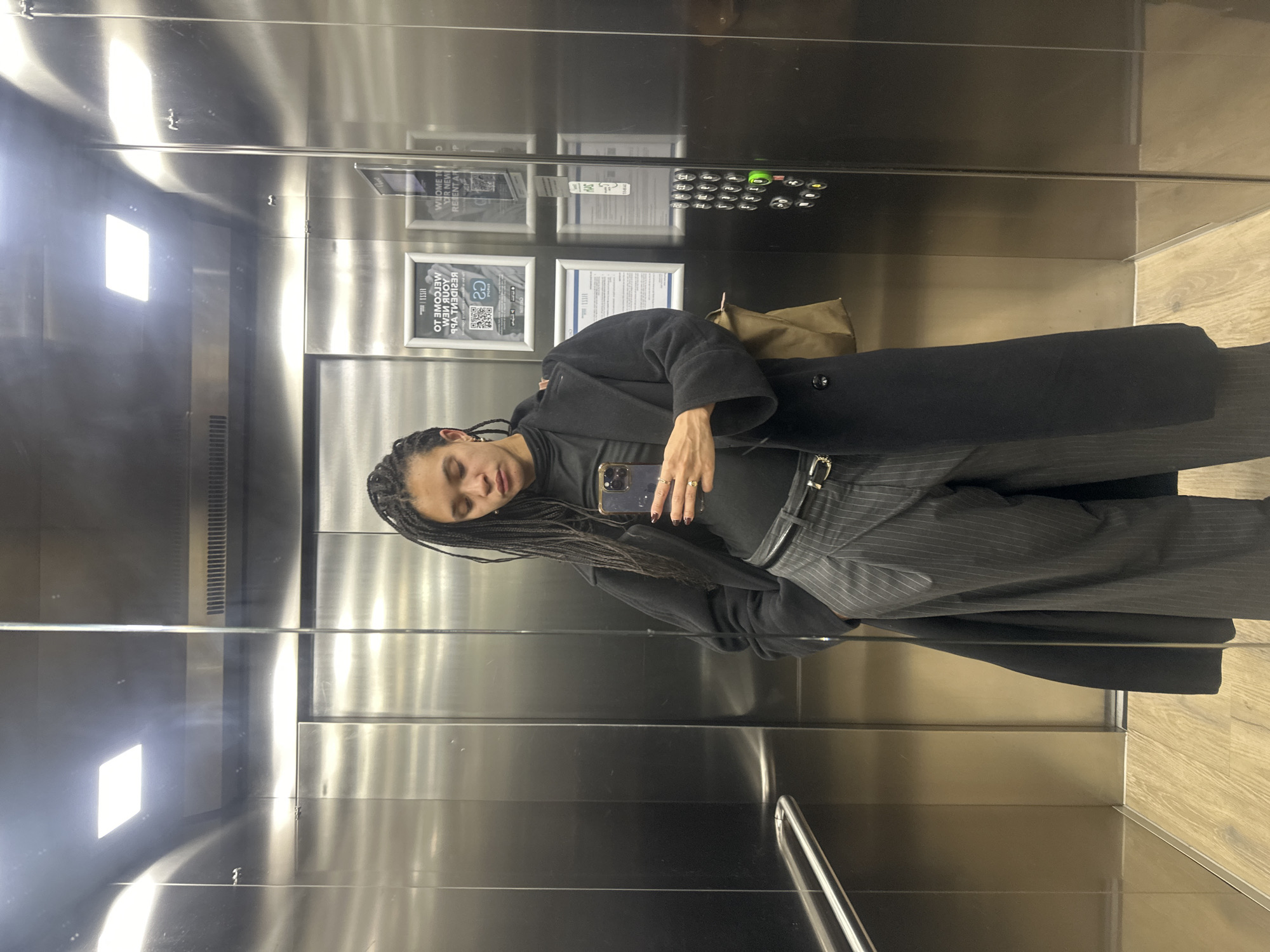 A woman with braided hair is taking a selfie in an elevator with stainless steel walls. She is wearing a black turtleneck, grey pinstripe pants with a black belt, and a black coat. She has rings on her fingers and is holding a smartphone. The elevator has a panel of buttons on the right and two framed signs. The floor of the elevator is wood-patterned. The reflection in the stainless steel walls provides further visual information about the elevator's interior. There is no visual evidence in the image to indicate that this location is in London, United Kingdom.