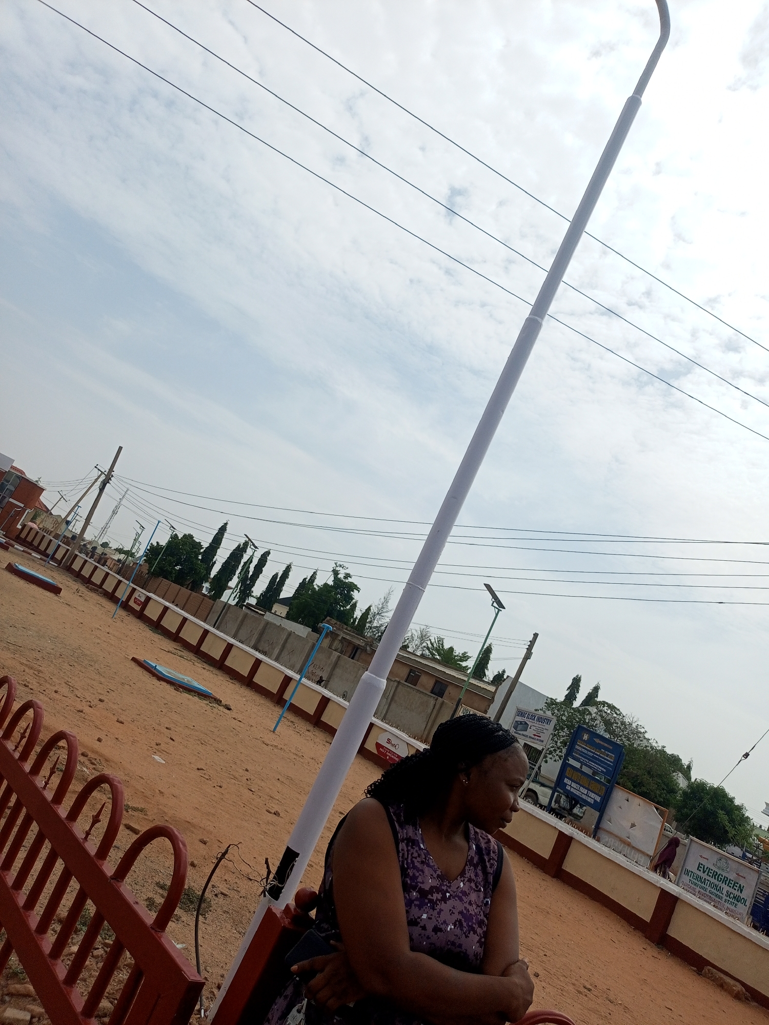 The image depicts a woman standing in an open, dusty area with a red metal fence in the foreground. Behind her is a tall, white lamppost. In the background, there is a low wall, behind which are buildings, trees, and signage, including a sign for "EVERGREEN INTERNATIONAL SCHOOL, TUMFURE GOMBE STATE." Other signs are visible, including one for "SEMAG BLOCK INDUSTRY" and a "NO SMOKING" sign. Overhead, electrical wires crisscross the sky, which is partly cloudy. The ground appears to be a mix of sand and dirt. The location is identified as Tumfure, Gombe State, Nigeria, based on the sign for Evergreen International School.