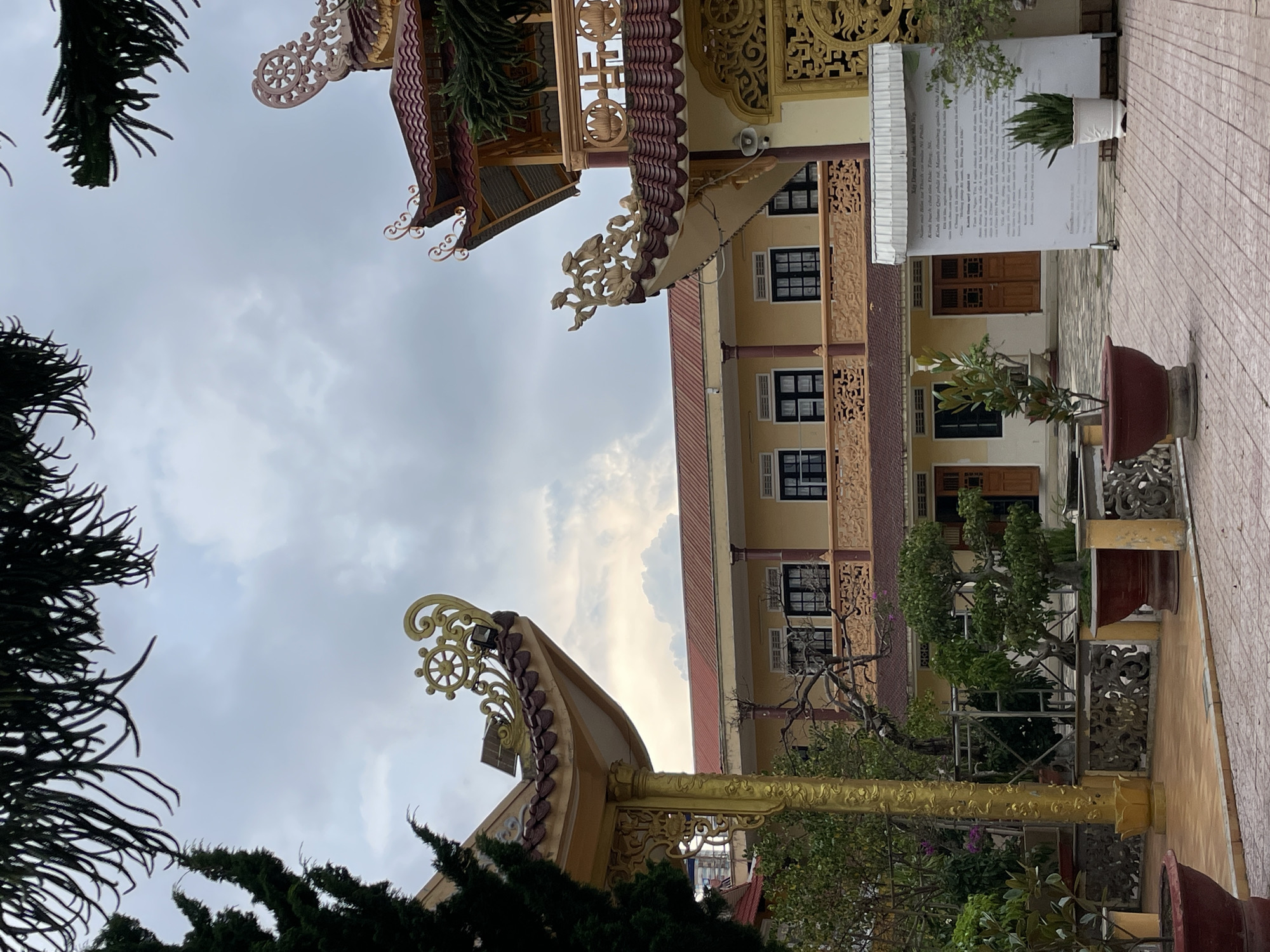 This image captures an outdoor scene at a Buddhist temple in Phường Xuân Hương, Đà Lạt, Vietnam. The foreground features a paved courtyard with red-tiled walkways. Several potted plants, including bonsai trees and snake plants, are strategically placed around the courtyard. Ornate, yellow-colored decorative pillars and railings add to the temple's aesthetic. In the mid-ground, a two-story building with a yellow facade, red-tiled roof, and dark-framed windows stands prominently. The building is framed by the eaves of the temple's more elaborate structures, characterized by red tiles, intricate golden decorative elements, and ornate roof designs, including a Dharma wheel symbol. The background is dominated by a cloudy sky. A white informational board with Vietnamese text is visible on the right side of the frame, near the entrance of the yellow building. No people are visible in the image.
