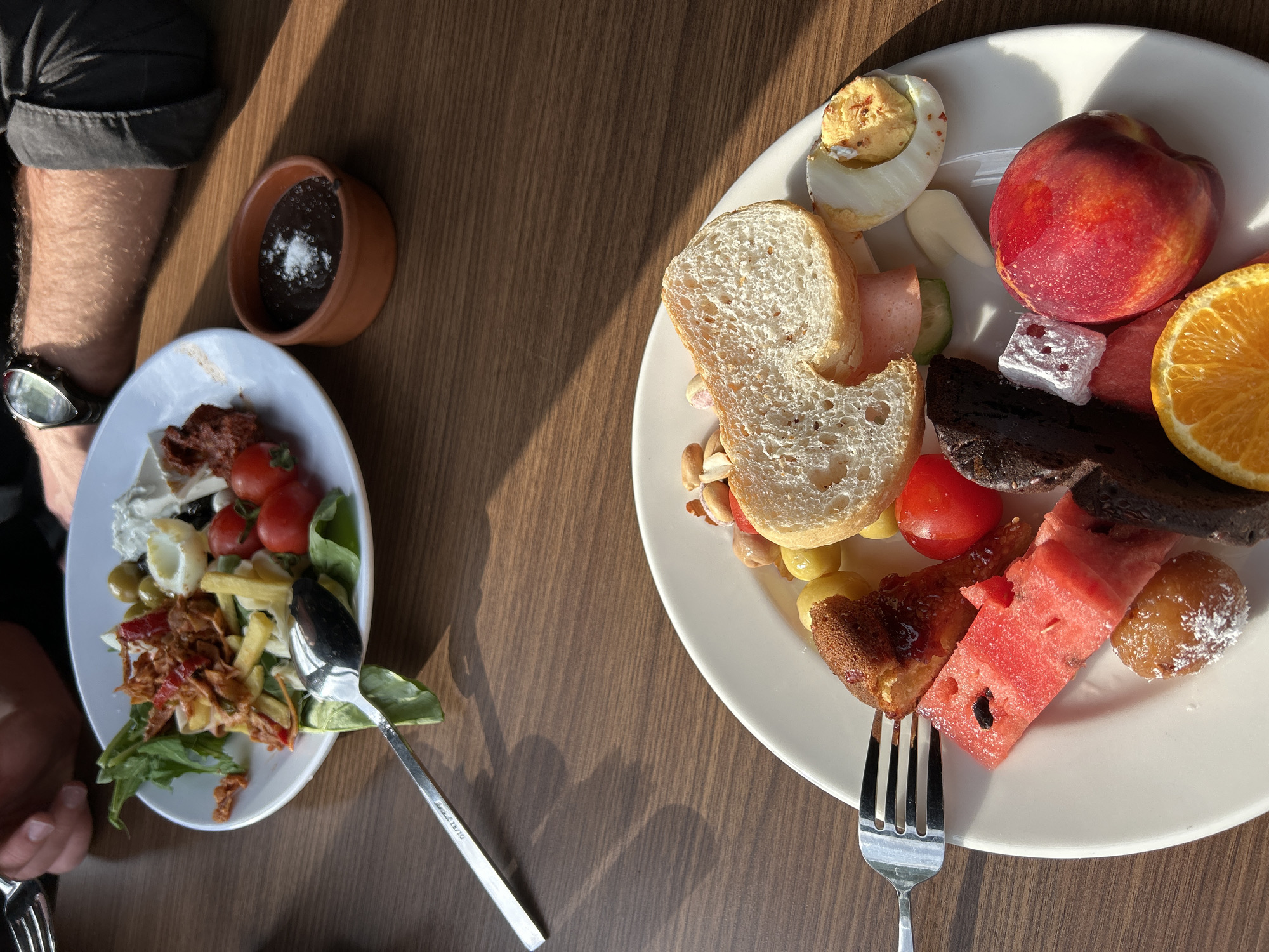 This overhead shot displays two plates of food laid out on a wooden table, likely for breakfast or brunch. The scene is illuminated by strong, direct sunlight, casting distinct shadows and indicating a morning or late afternoon setting on a sunny day. The location is given as Esenler, Turkey.
On the left, a person's right arm is visible, wearing a dark shirt with rolled-up sleeves and a silver watch. Their hand holds a fork. Their oval plate features a vibrant salad composed of green leaves, cherry tomatoes, olives, white cheese or cream, what appears to be fried calamari, and a topping of shredded red-orange peppers or relish. Adjacent to this plate is a small, brown terracotta bowl filled with a dark, pudding-like substance, sprinkled with white powder. A silver spoon, bearing the text "OLIVES 18/10," rests near the salad.
The round plate on the right, for a second diner, presents a diverse array of items. It includes two slices of white bread, half a hard-boiled egg seasoned with spices, a whole red-orange nectarine or peach, a slice of orange, several pieces of watermelon, a dark slice of cake or brownie, a white cubed sweet resembling Turkish delight, and a yellowish pastry slice. Other accompaniments include small nuts or dried fruits, cucumber slices, and a piece of pink ham or salami. A fork is positioned under the watermelon. The variety of fresh fruit, bread, eggs, and sweet items is typical of a large breakfast spread, often found in Mediterranean or Middle Eastern regions.