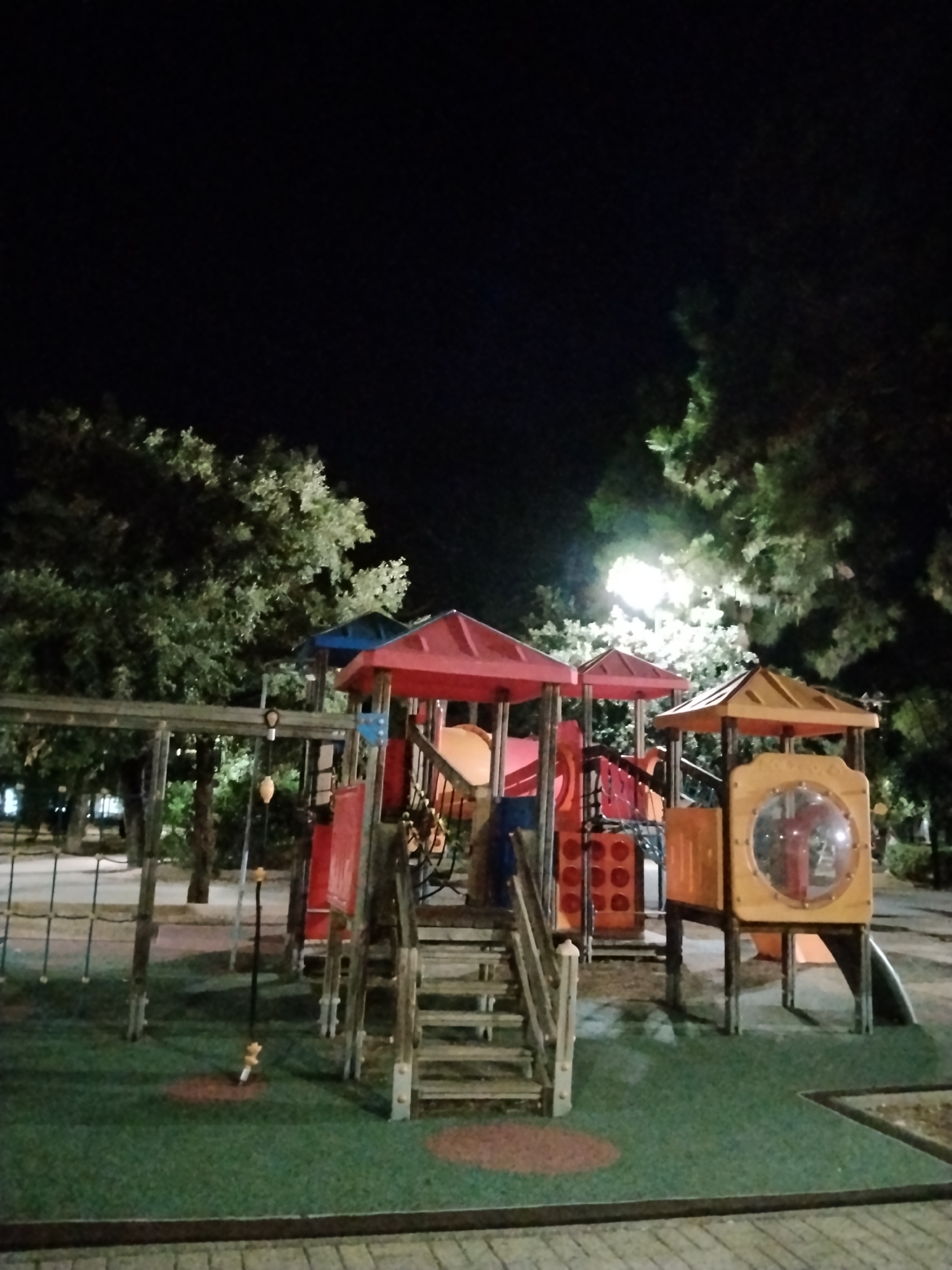 The image shows a playground at night in Bitonto, Italy. The playground features a multi-colored play structure with red and yellow roofs, slides, and climbing elements. A wooden ladder leads up to one of the platforms. A net climbing structure is visible to the left, and a swing hangs from a wooden beam. The ground is covered with green, soft surfacing, with circular red pads. Trees surround the playground, and a bright street light illuminates the scene. The sky is dark.