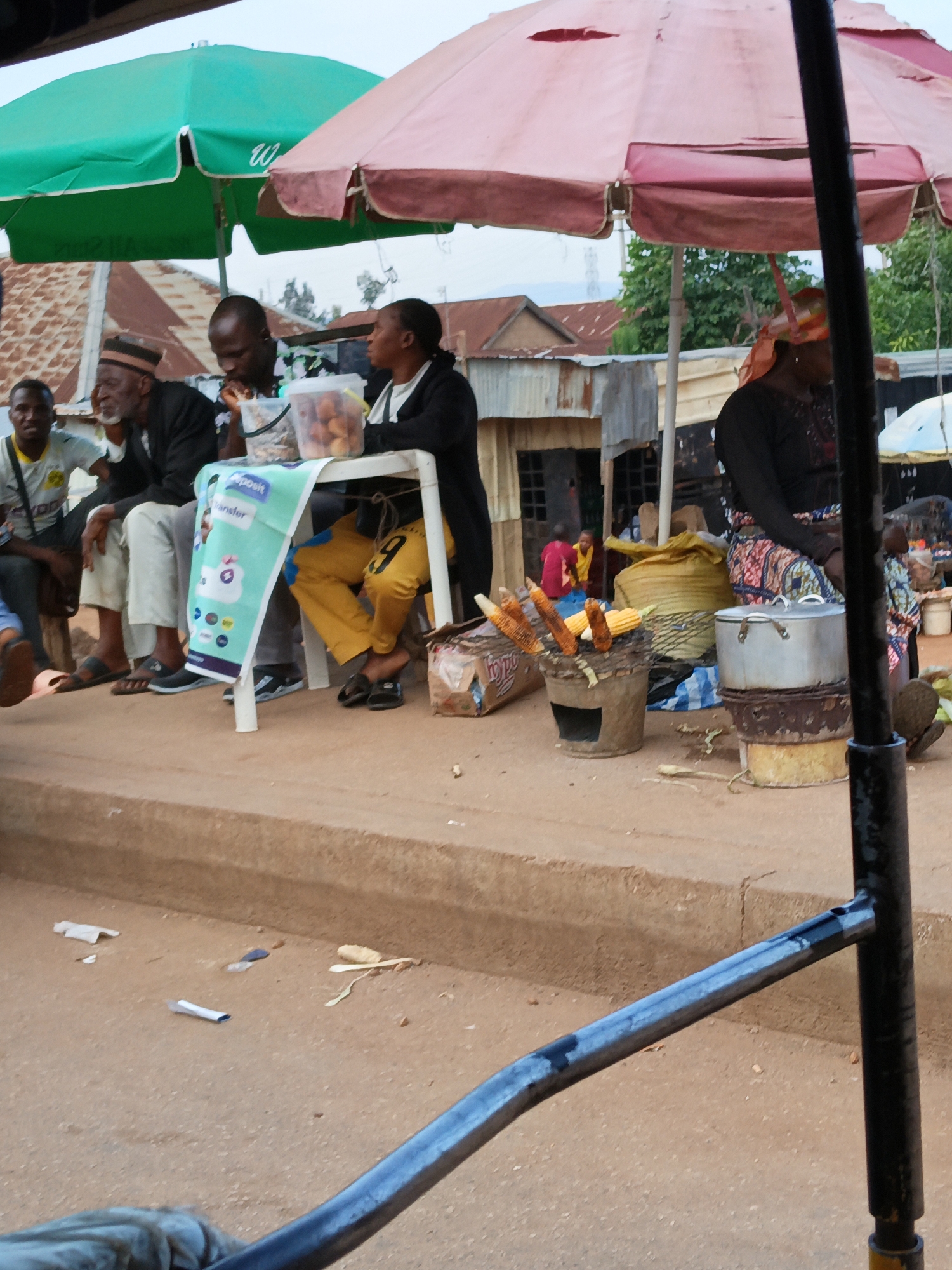In Bukuru, Nigeria, an outdoor market scene unfolds under the shade of two umbrellas, one green and one pink. Several individuals are present, some seated and others standing.
In the foreground, a metal railing, partially obscured by the camera's perspective, separates the viewer from the scene. To the right of the railing, a black metal pole stands vertically, supporting the structure of the scene.
In the center of the image, a white plastic table is set up, draped with a colorful banner advertising "Deposit Transfer." On the table, a clear plastic container holds several fried dough balls, resembling puff puffs.
Several individuals are gathered around the table. On the left, a man wearing a black cap and traditional attire is seated, looking downwards. To his right, another man in a black jacket is also seated, eating. Further to the right, a person in yellow pants is seated, their legs crossed, facing away from the viewer.
To the right of the table, a woman wearing a patterned skirt and headscarf is seated. In front of her, a collection of corn on the cob is being roasted over a makeshift grill, a cylindrical metal container with an opening at the bottom. Beside the grill, a large silver pot sits on a stacked base. Several large bags, possibly filled with produce or grains, are visible behind the cooking area.
In the background, glimpses of market stalls with corrugated iron roofs and wooden structures suggest the bustling environment of a Nigerian market. The overall scene depicts a snapshot of daily life and commerce in Bukuru.