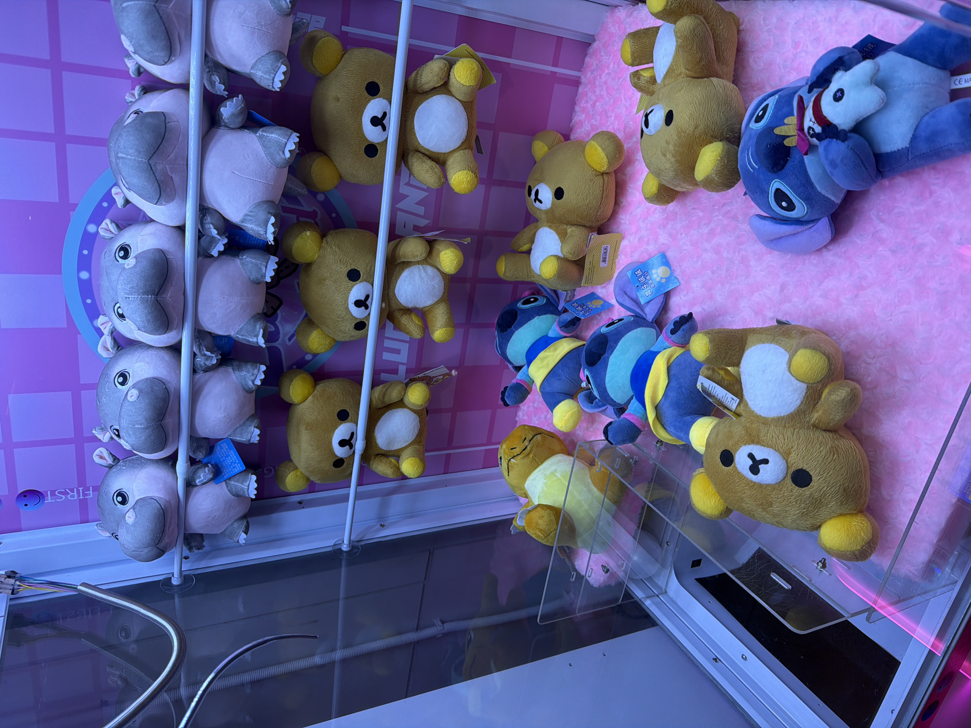 This image captures the interior of a claw game machine located in an indoor commercial establishment in Bangkok, Thailand. The machine features a transparent front panel and an internal pink wall with a subtle grid pattern and partial white text reading "FIRST". The bottom surface of the play area is covered with a fluffy pink fabric.
Arranged within the machine are numerous plush toys:
* The top row displays six light grey plush hippopotami, each with a white belly, light pink inner ears, and black eyes, suspended by their backs. A blue tag is attached to one.
* The middle row holds three light brown plush bears, resembling Rilakkuma, with white bellies, yellow ears, and black embroidered faces, also suspended by their backs. A white tag is visible on the rightmost bear.
* On the bottom pink-lined platform, at least four additional light brown Rilakkuma-like plush bears are scattered in various prone positions. One yellow plush character, resembling Pikachu, is also present on its back on the left side of the platform.
* Two blue plush characters, resembling Stitch from Disney's Lilo & Stitch, are lying on their backs. The Stitch on the right is holding a smaller white plush toy with a red accessory. Tags are visible near these plush toys, one with blue text.
A metallic claw mechanism, connected by multi-colored wires, extends from the upper left into the machine. A clear plastic prize chute with a dark opening is visible in the lower left section of the machine. The scene is illuminated by cool-toned artificial lighting. No human figures are present.