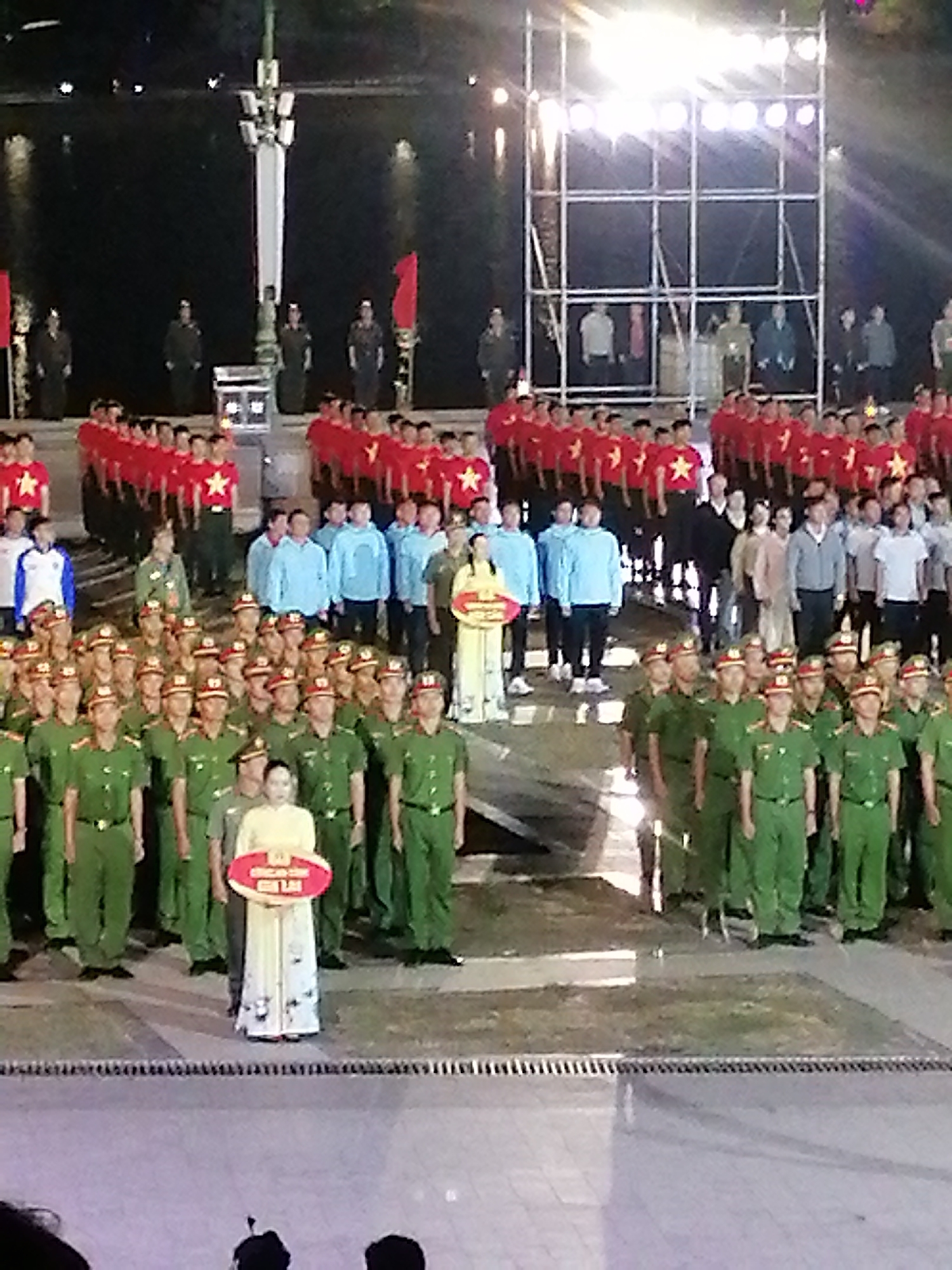 A group of people in military uniforms stand at attention in what appears to be an outdoor public space at night. The people are arranged in rows, with several individuals in the foreground wearing green uniforms. A woman in a pale yellow dress stands in the center of the front row, holding a red oval sign with white text. Behind the uniformed group, another group of people is visible, some wearing red shirts with yellow stars, and others in light blue tops. In the background, there is a body of water and a tall structure with lights, suggesting a stage or lighting rig. The overall setting suggests an organized event, possibly a ceremony or performance, taking place outdoors after dark. The lighting is bright, indicating artificial illumination. The scene is likely in Da Lat, Vietnam, based on the provided location context. The mood appears formal and organized.