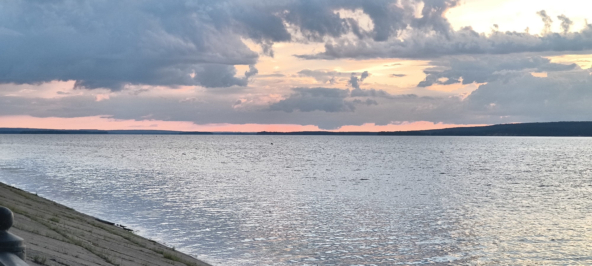 The image depicts a wide, calm body of water, likely a lake or a large river, under a dramatic sky filled with clouds. The water's surface is rippled, reflecting the soft colors of the sunset or sunrise. A sloping concrete embankment lines the left foreground of the image, with some sparse greenery growing between the slabs. A dark, rounded object, possibly a railing post, is visible on the far left. The horizon is marked by a distant, tree-covered shoreline. The sky is a canvas of soft pinks, oranges, and grays, with darker, more turbulent clouds dominating the upper portion of the frame. The overall impression is one of natural beauty and tranquility. The location is not specified within the city of Unknown, Unknown.
